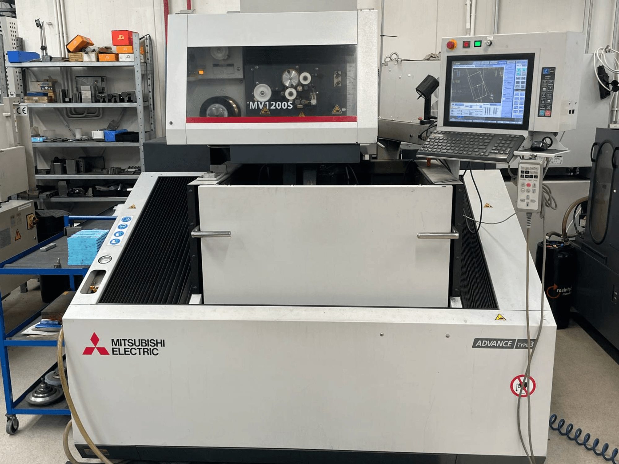 „Mitsubishi MV1200S“ vielinės elektros erozijos staklių vaizdas iš priekio, kuriame matomas valdymo pultas ir darbo zona, taip pat matomi saugos įspėjimai.