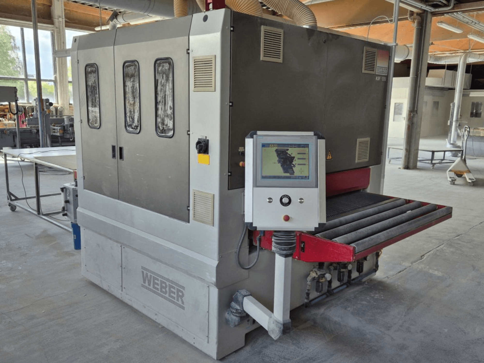 WEBER KSF-3-1350 juostinis šlifuoklis, šoninis vaizdas, kuriame matyti valdymo įtaisai, maitinimo skydelis ir šlifavimo konvejeris. Pramoninė aplinka su matomomis mašinomis.