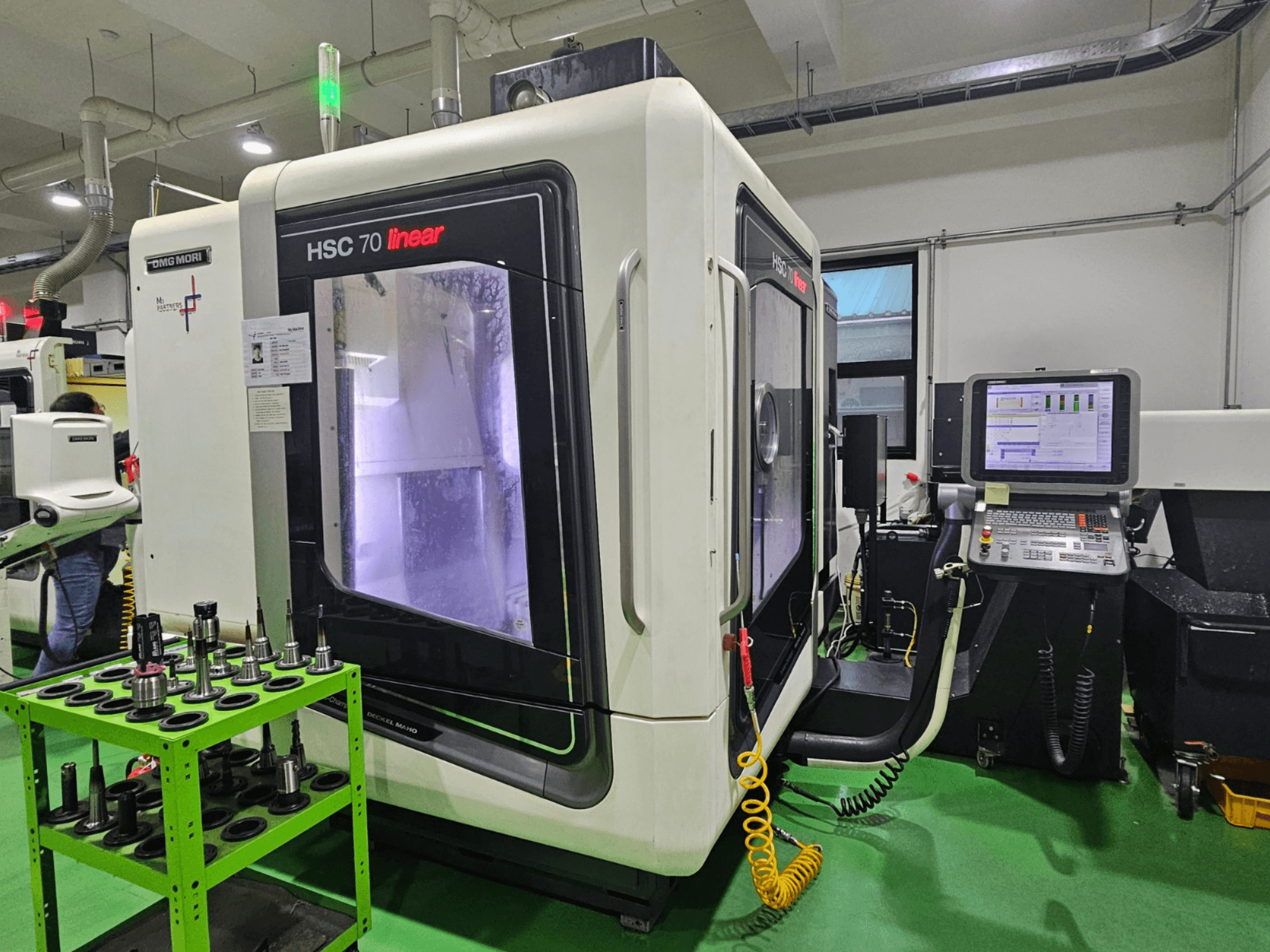 DMG MORI HSC 70 linijinis universalusis apdirbimo centras, vaizdas iš priekio, kai ant žalio stalo yra įrankių laikikliai ir matomas valdymo skydelis.