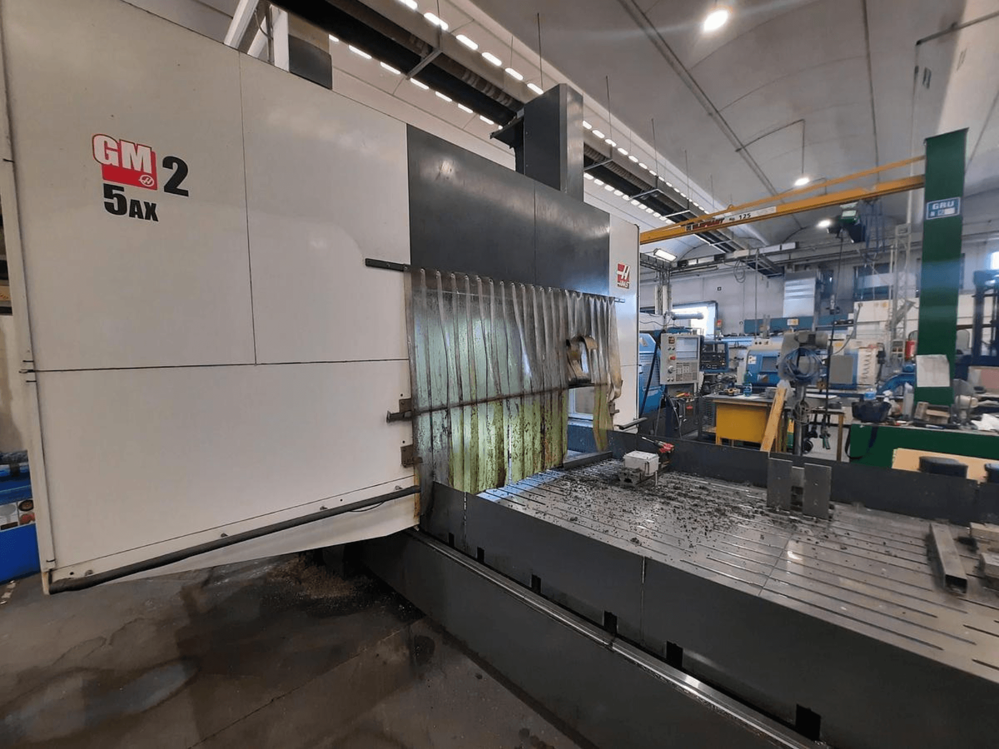 GM 2 5AX CNC staklės dirbtuvėse, šoninis vaizdas, kuriame matoma darbinė zona ir metalo drožlės ant pado paviršiaus.