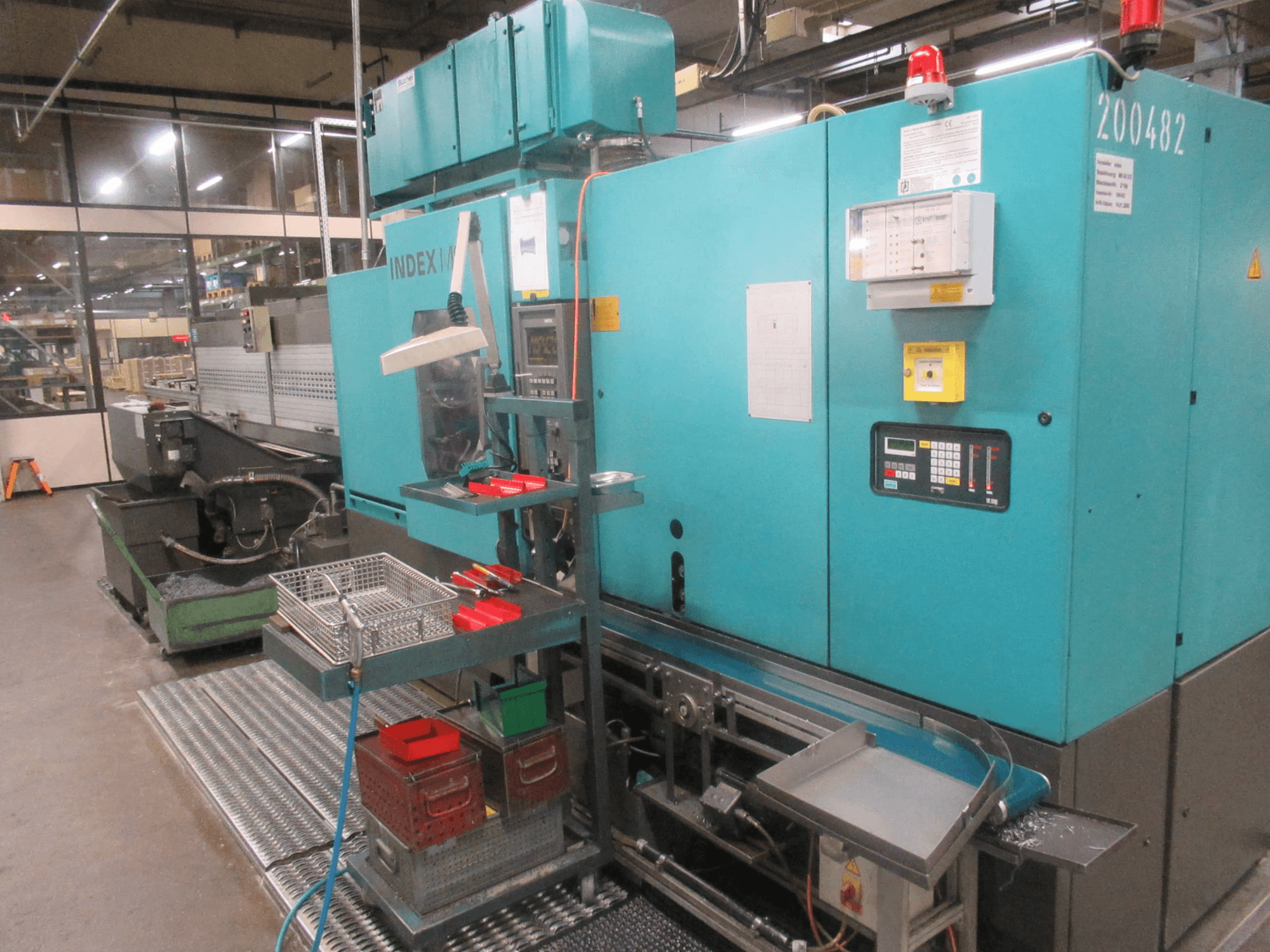 Turkio spalvos INDEX staklės gamykloje, vaizdas iš šono; yra valdymo pultas, įrankių saugykla ir konvejeris. Matomos talpyklos, žarnos ir signalizacija.