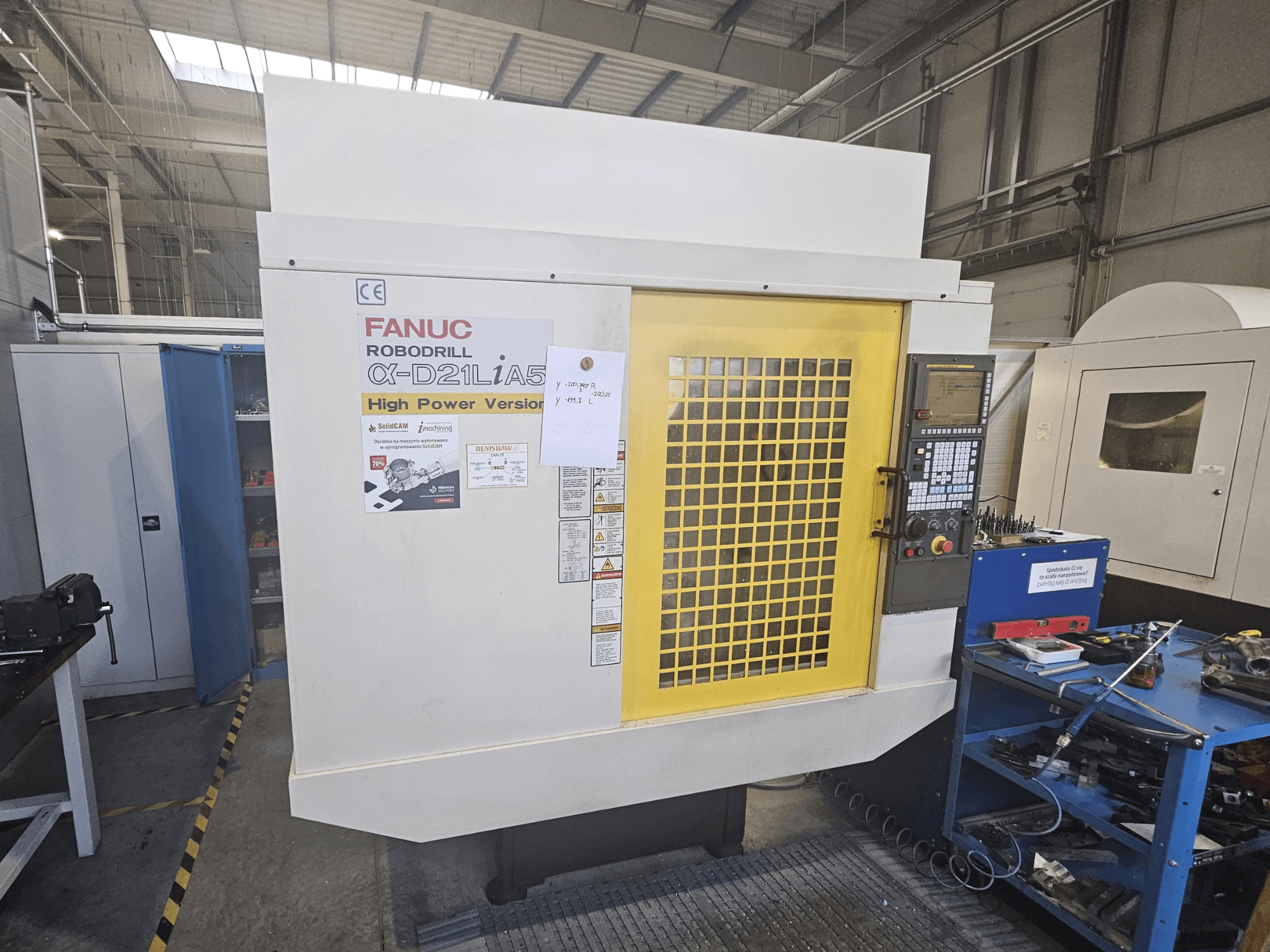 FANUC Robodrill α-D21LiA5 priekinis vaizdas su geltonomis apsauginėmis durelėmis, valdymo skydeliu ir šalia esančiais darbo vietos įrankiais.