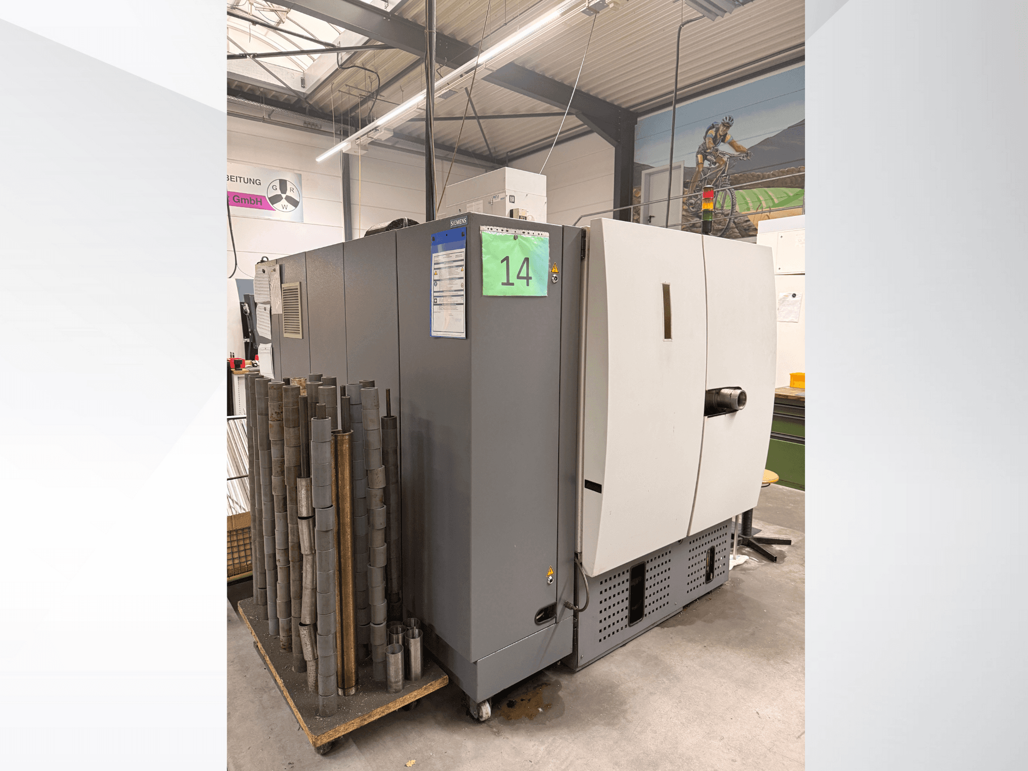 Pilkos "Siemens" pramoninės staklės, vaizdas iš šono, šalia jų - cilindrinių metalo gabalų atsargos ir matomos įrangos etiketės.