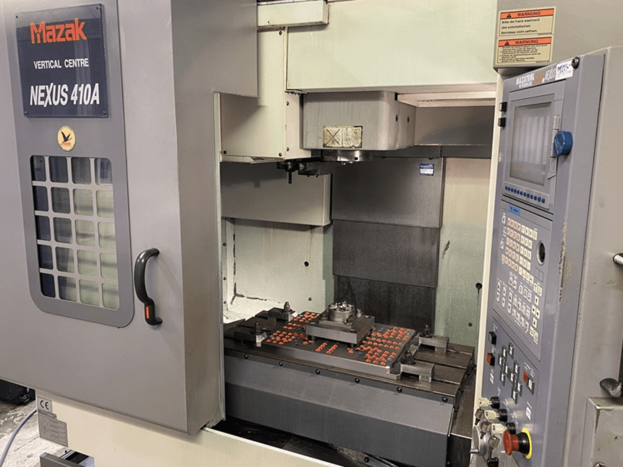 Vertikalaus apdirbimo centras Mazak VCN410, priekinis vaizdas, kuriame matomas valdymo skydelis ir staklių vidus su pritvirtinta apdirbama detale.