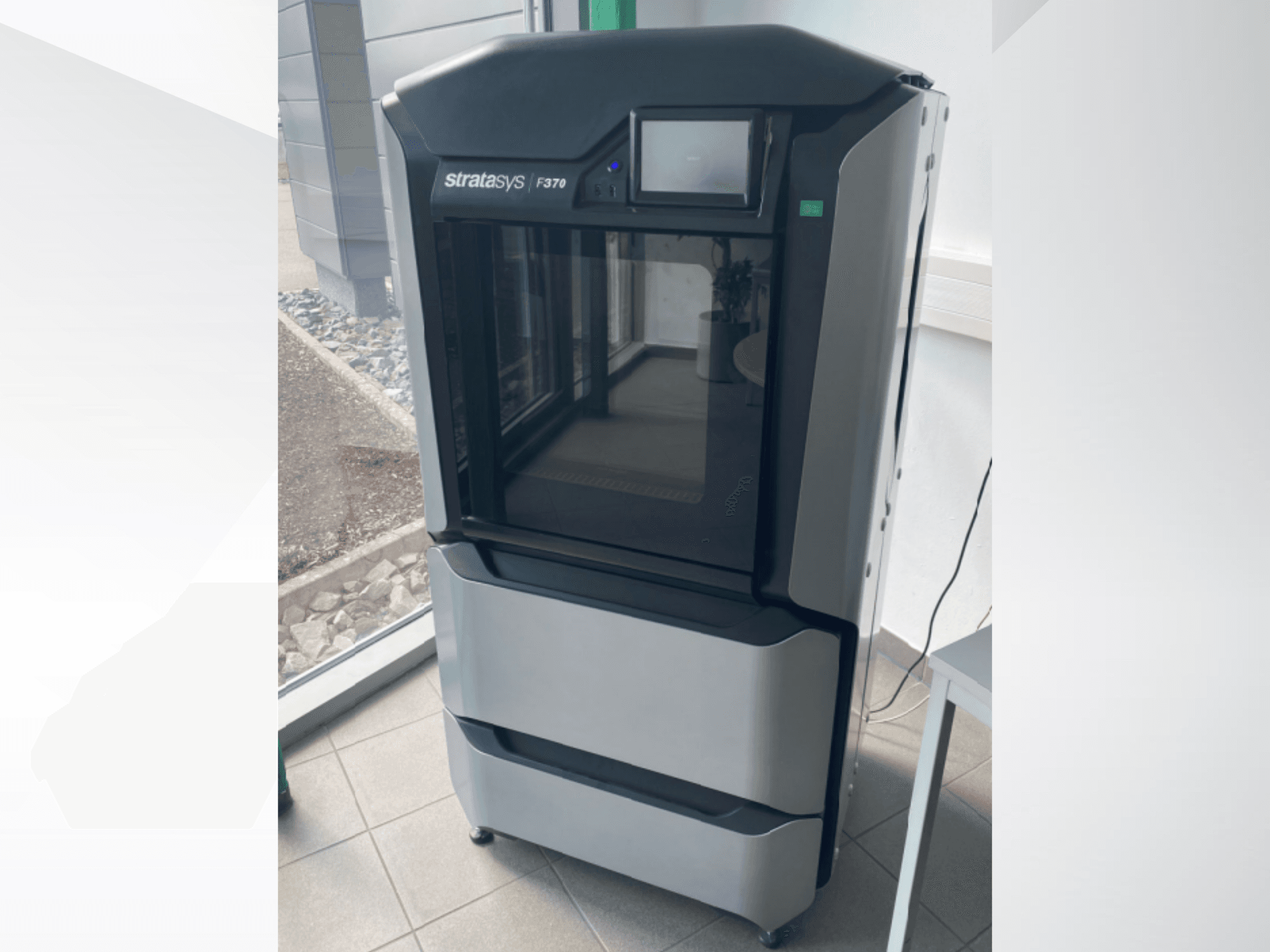 Plastikinis 3D spausdintuvas "Stratasys F370", vaizdas iš priekio, demonstruojantis elegantišką juodai pilką dizainą su jutikliniu ekranu.