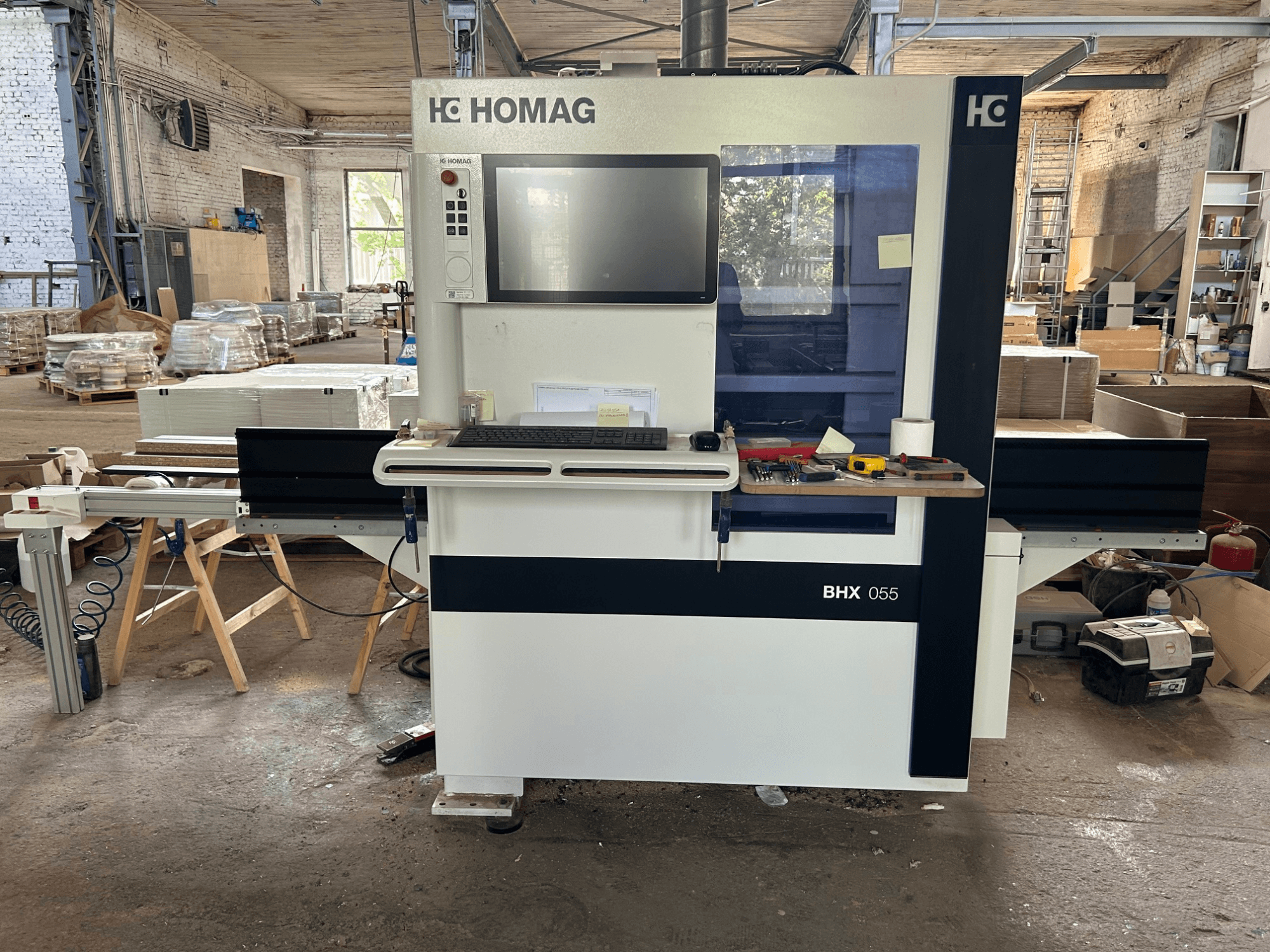 HOMAG WEEKE OPTIMAT BHX 055 CNC apdirbimo centras, vaizdas iš priekio, kuriame matomas valdymo pultas ir darbo zona su įrankiais.