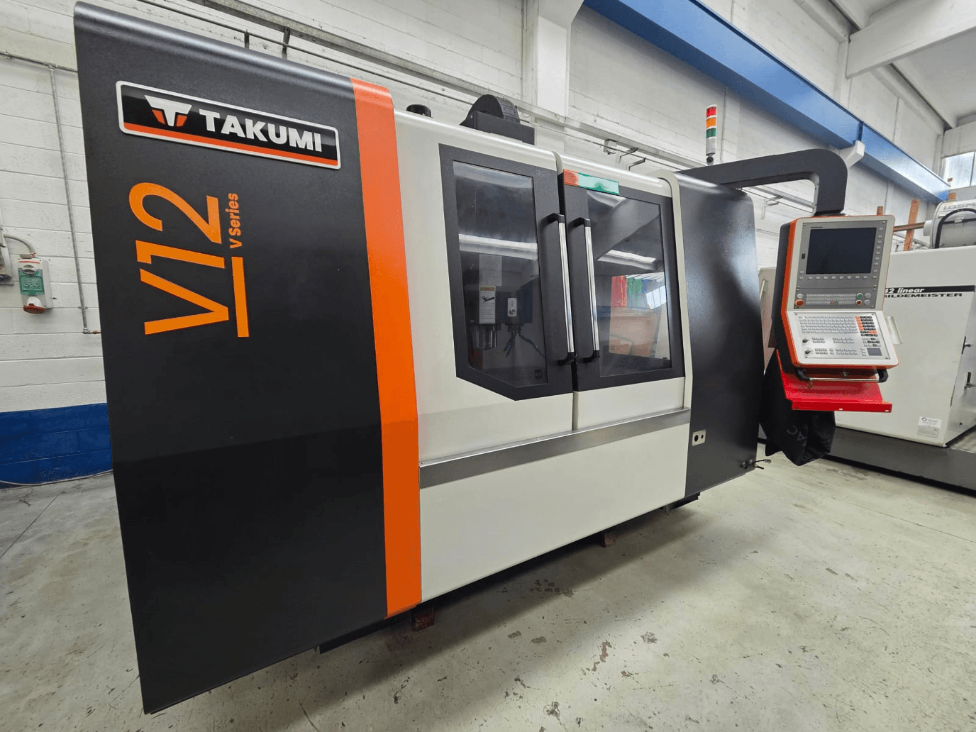 Takumi V12 V serijos CNC frezavimo staklės, vaizdas iš priekio, su valdymo pultu ir dideliais stikliniais langais.