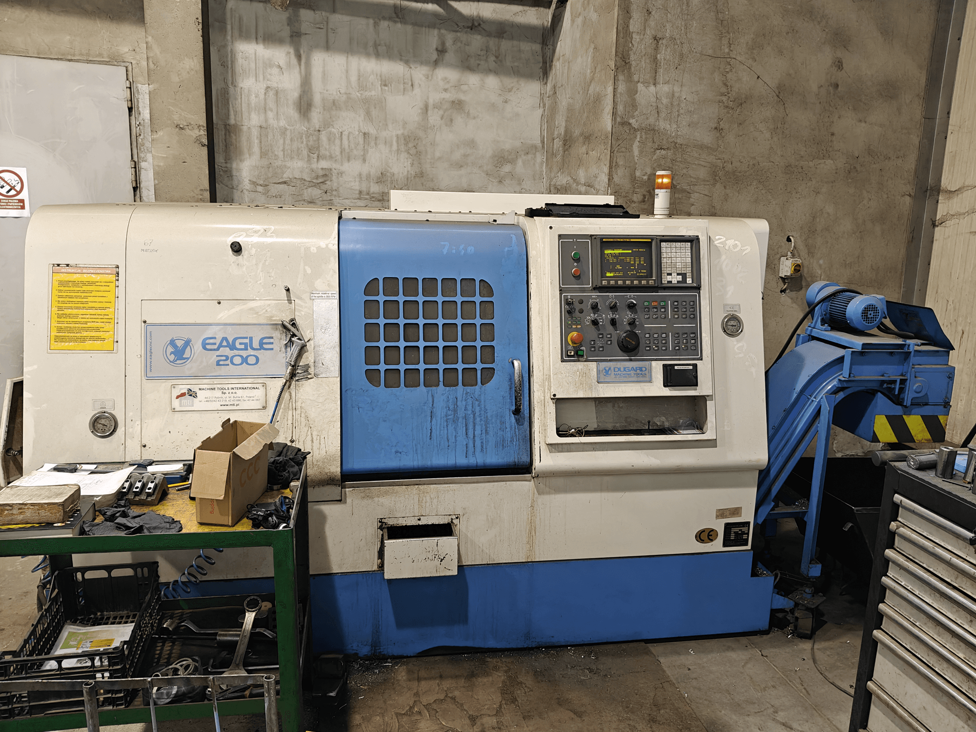 CNC staklės "Eagle 200", vaizdas iš priekio, valdymo skydelis, įvairūs įrankiai ant žalio vežimėlio. Matomas nusidėvėjimas ir veikimo lemputės.