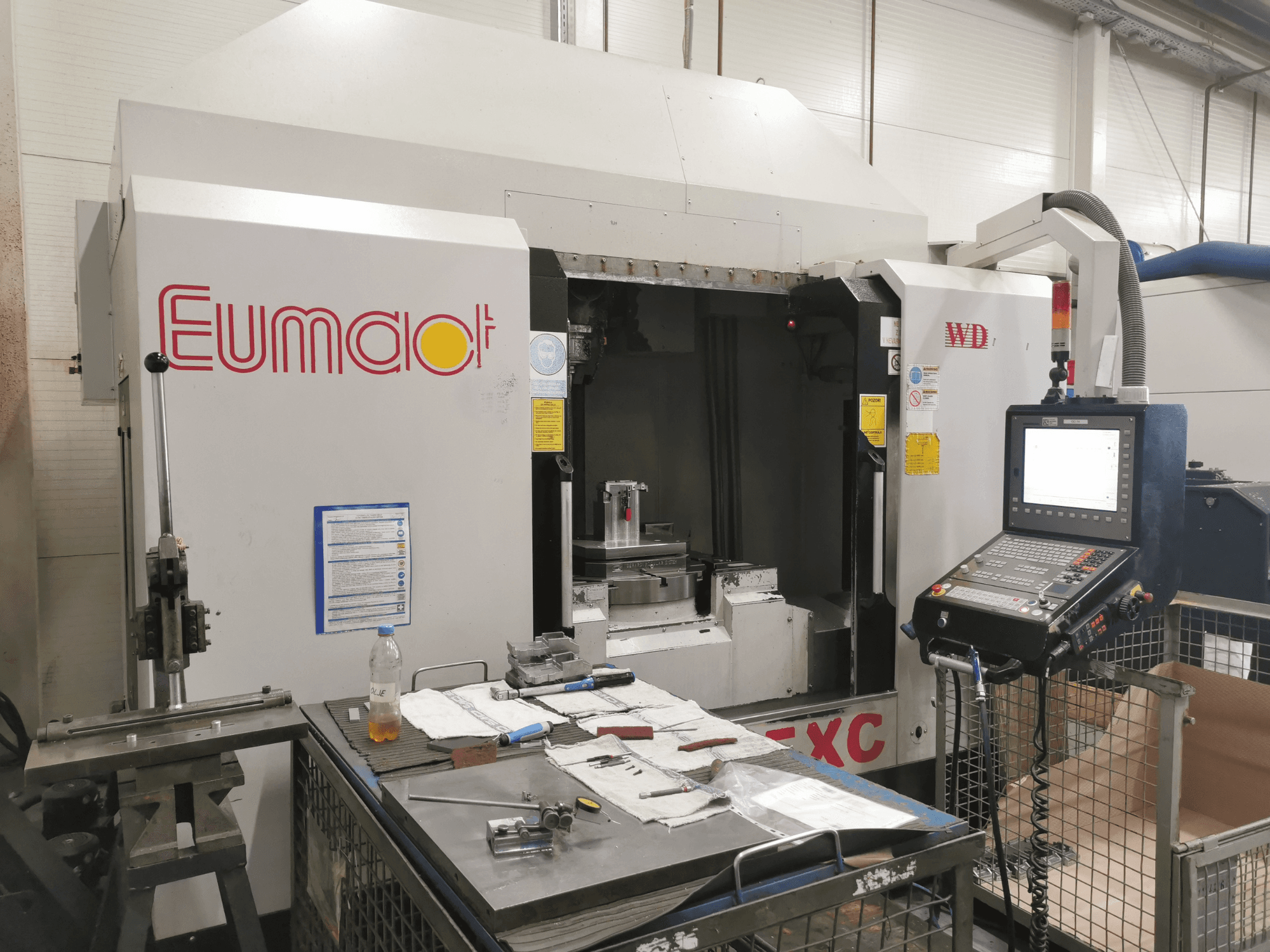 "Eumdaot" CNC staklių vaizdas iš priekio su valdymo skydeliu, įrankiais ir komponentais bei medžiagomis užgriozdintu darbo paviršiumi.