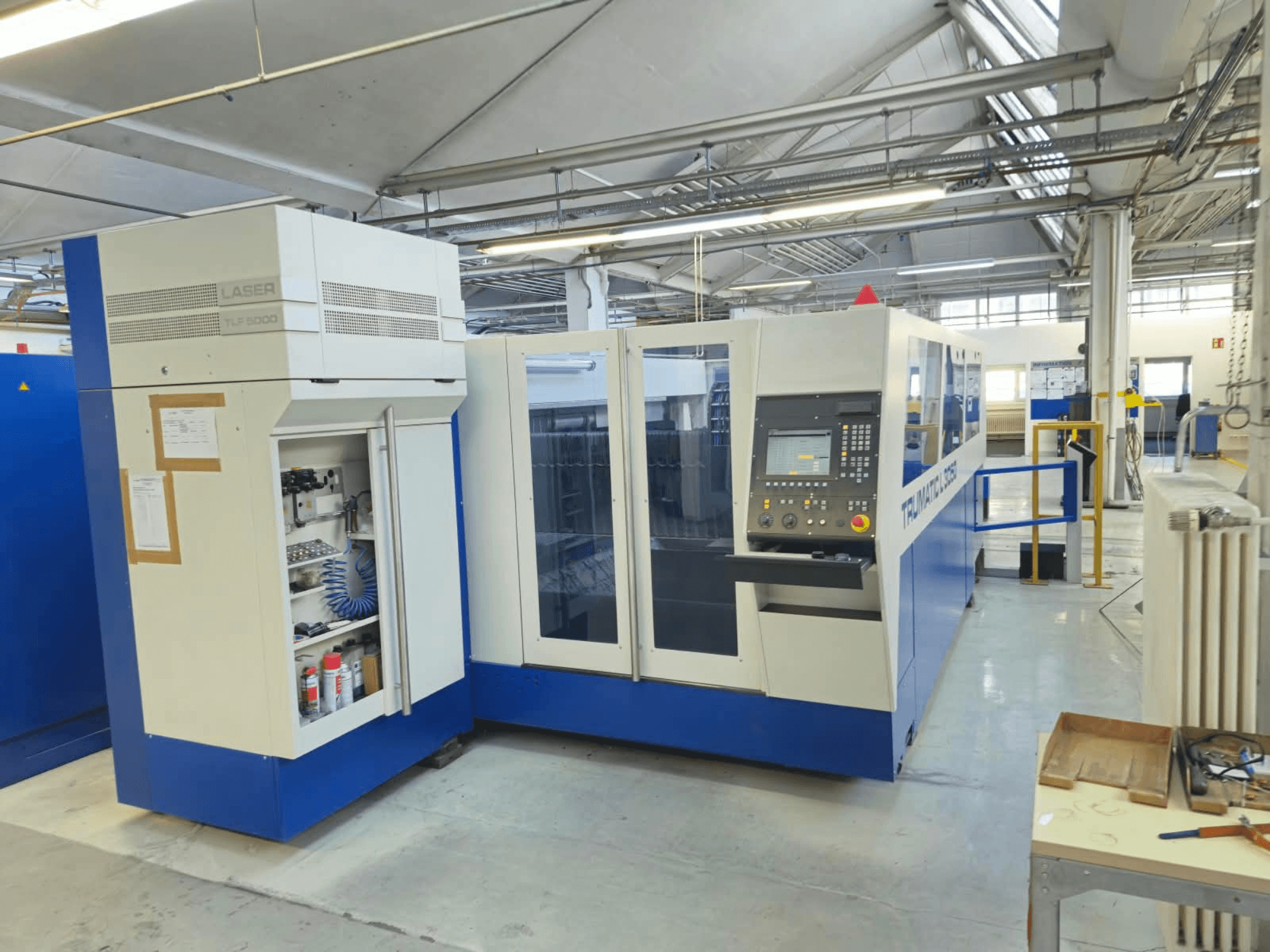 „TRUMPF TruLaser L3050“ CO₂ lazerio pjovimo staklės, vaizdas iš priekio, su valdymo skydeliu ir techninės priežiūros durelėmis, gamyklinėse sąlygose.