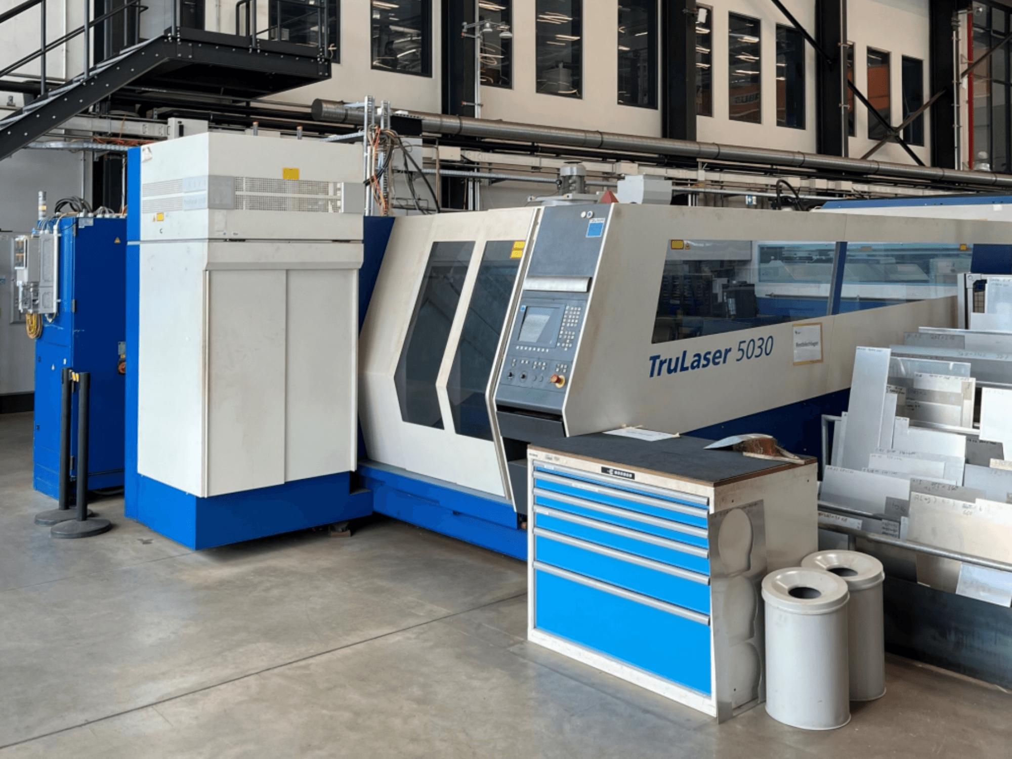 „TRUMPF TruLaser 5030“ CO₂ lazerinio pjovimo staklių vaizdas iš priekio, kuriame matomas valdymo pultas ir šalia esantis darbinis stalas su metaliniais lakštais.