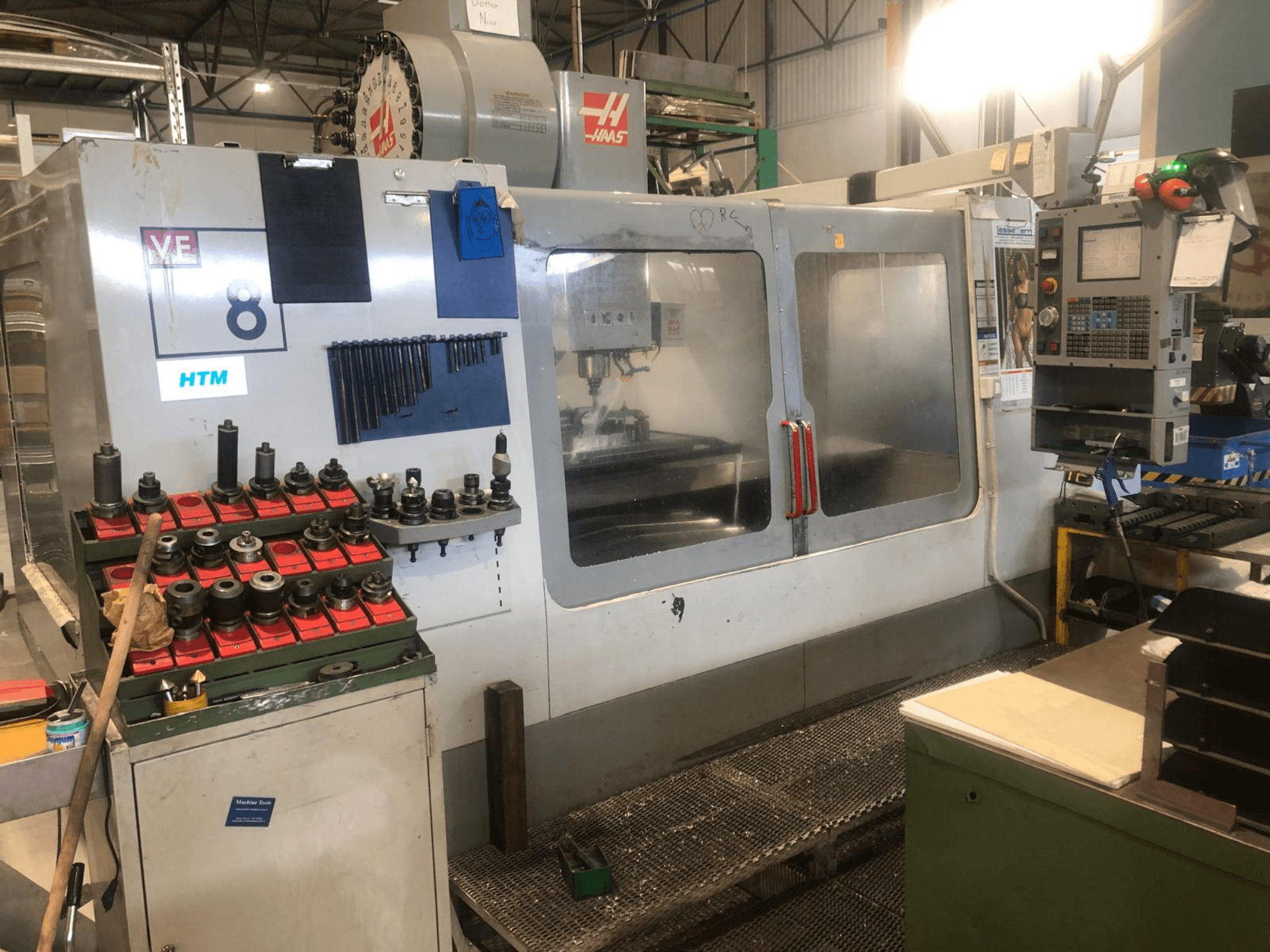 Haas CNC staklės dirbtuvėse, vaizdas iš priekio; rodomi įrankiai ir valdymo įtaisai, matomos modelio detalės ir sutvarkyti įrankių laikikliai.