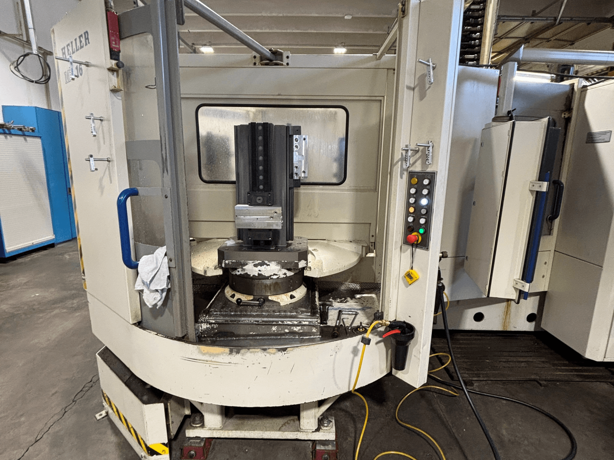 Vertikalaus modelio "Heller" apdirbimo centras, vertikalusis modelis, priekinis vaizdas, kuriame matomas suklys, valdymo skydelis ir stalas su pritvirtinta įranga.