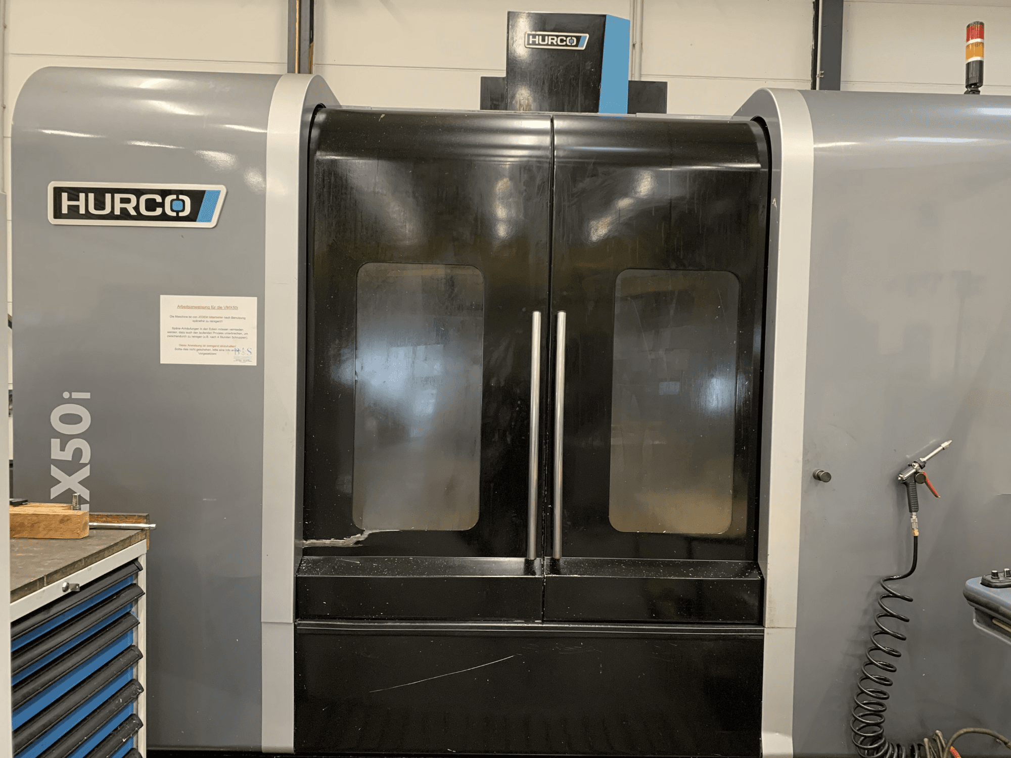 Hurco VX50i CNC staklių su dvejomis durelėmis vaizdas iš priekio, matomi techniniai valdikliai ir techninės priežiūros ženklas šone.