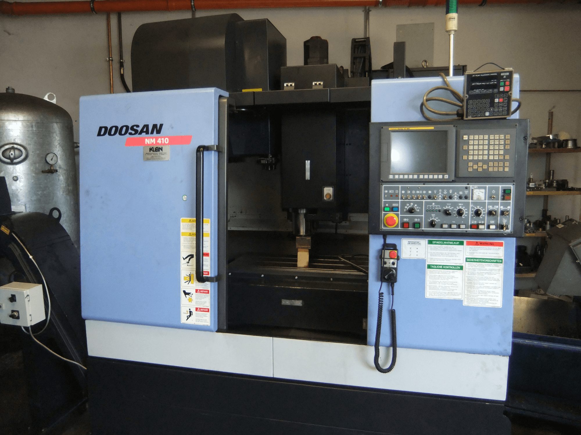 Vertikalusis apdirbimo centras „DOOSAN NM 410“, vaizdas iš priekio, kuriame matomas valdymo pultas, suklio zona ir saugos etiketės.