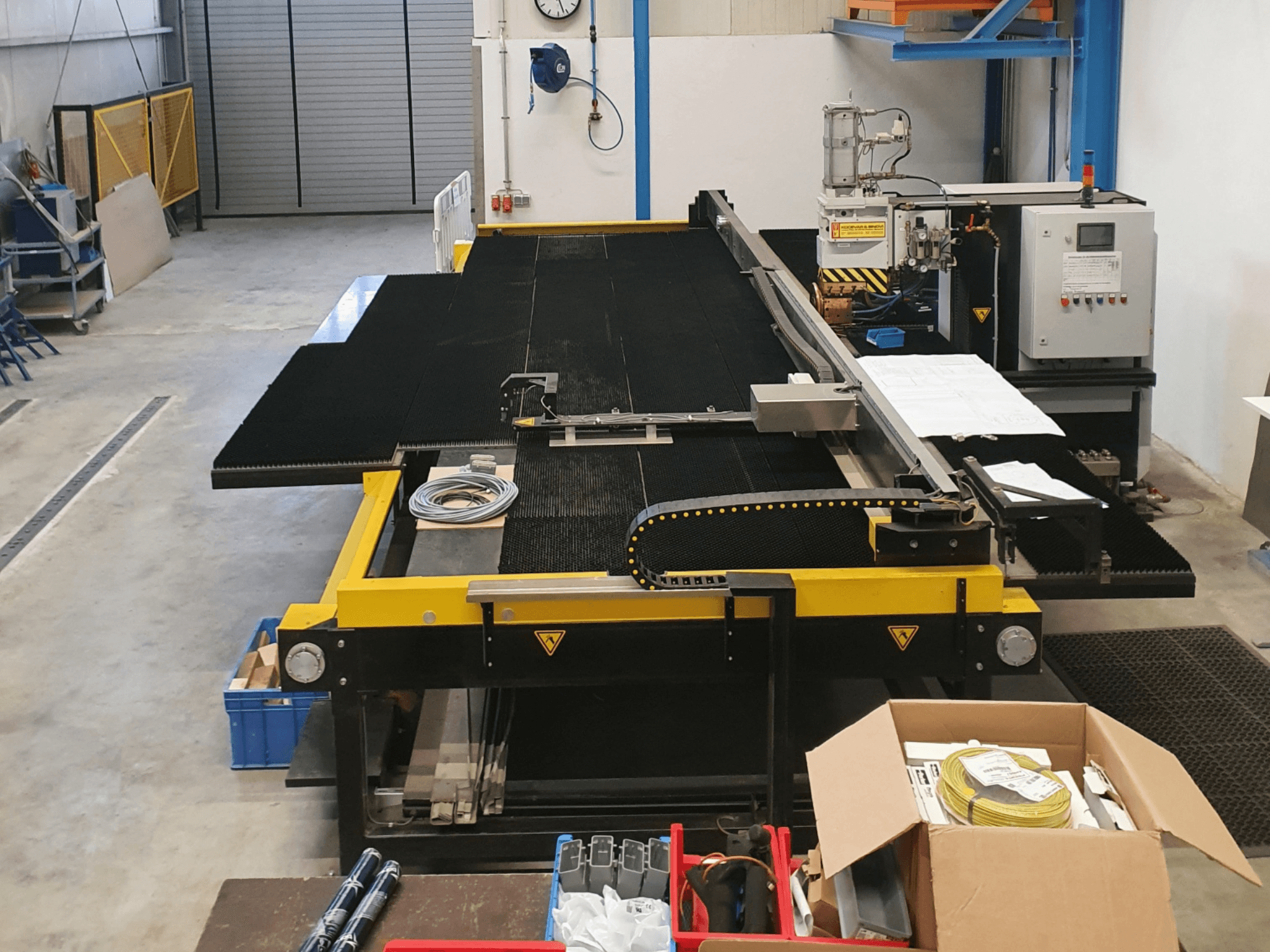 Geltonos ir juodos spalvos pramoninės pjaustymo staklės, vaizdas iš priekio, kuriame matomi gamykloje esantys valdikliai ir priedai.