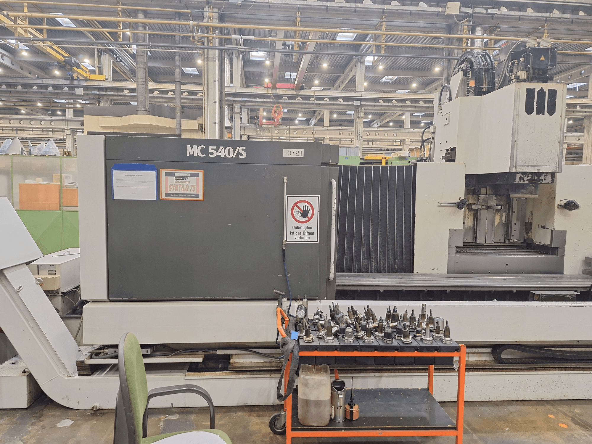 Vertikalusis apdirbimo centras STAMA MC 540S dirbtuvėse, šoninis vaizdas su įrankių laikikliais ant šalia esančio vežimėlio.