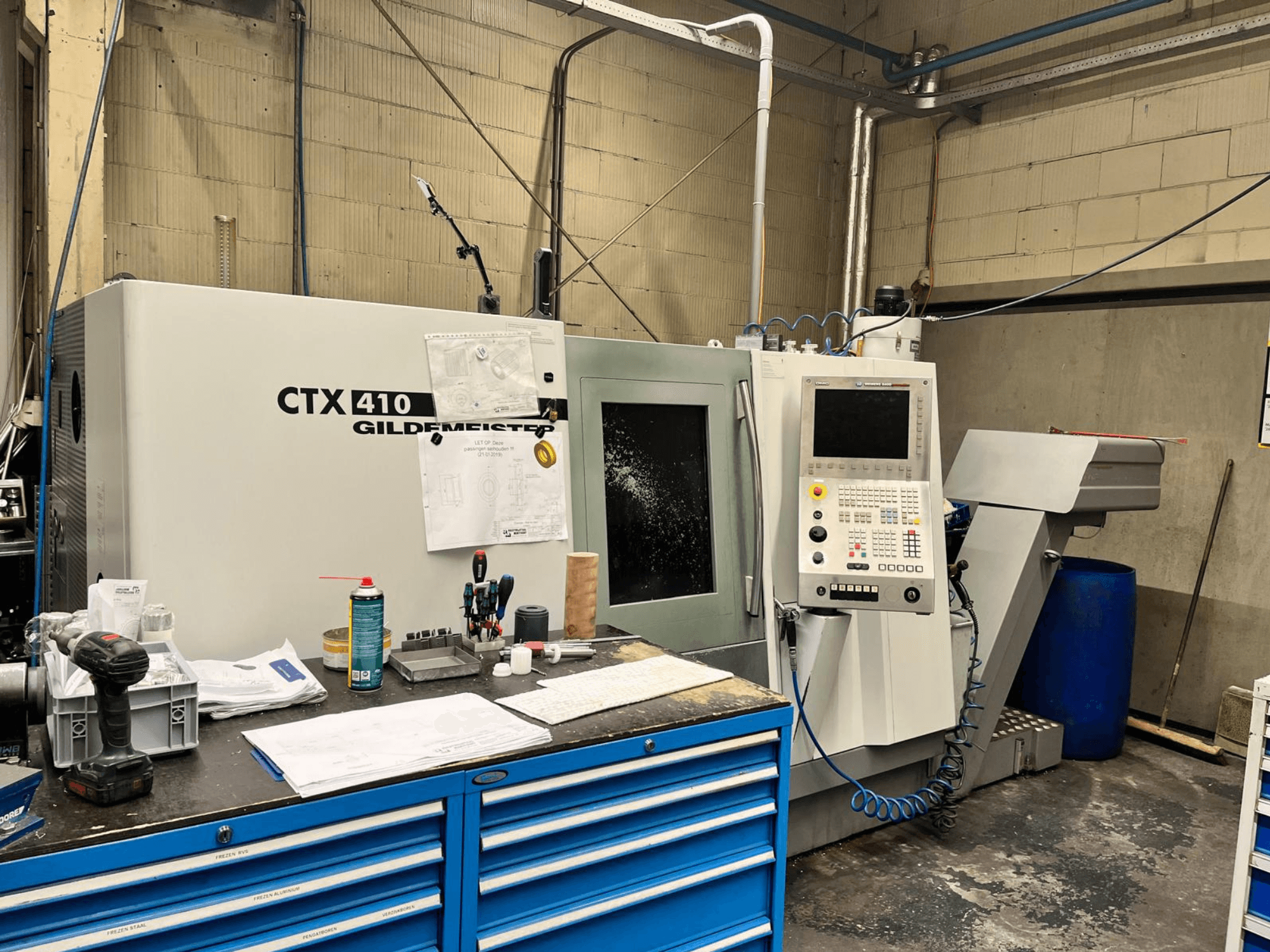 Gildemeister CTX 410 CNC staklės dirbtuvėse, vaizdas iš priekio, valdymo skydelis ir įvairūs įrankiai ant darbo stalo.