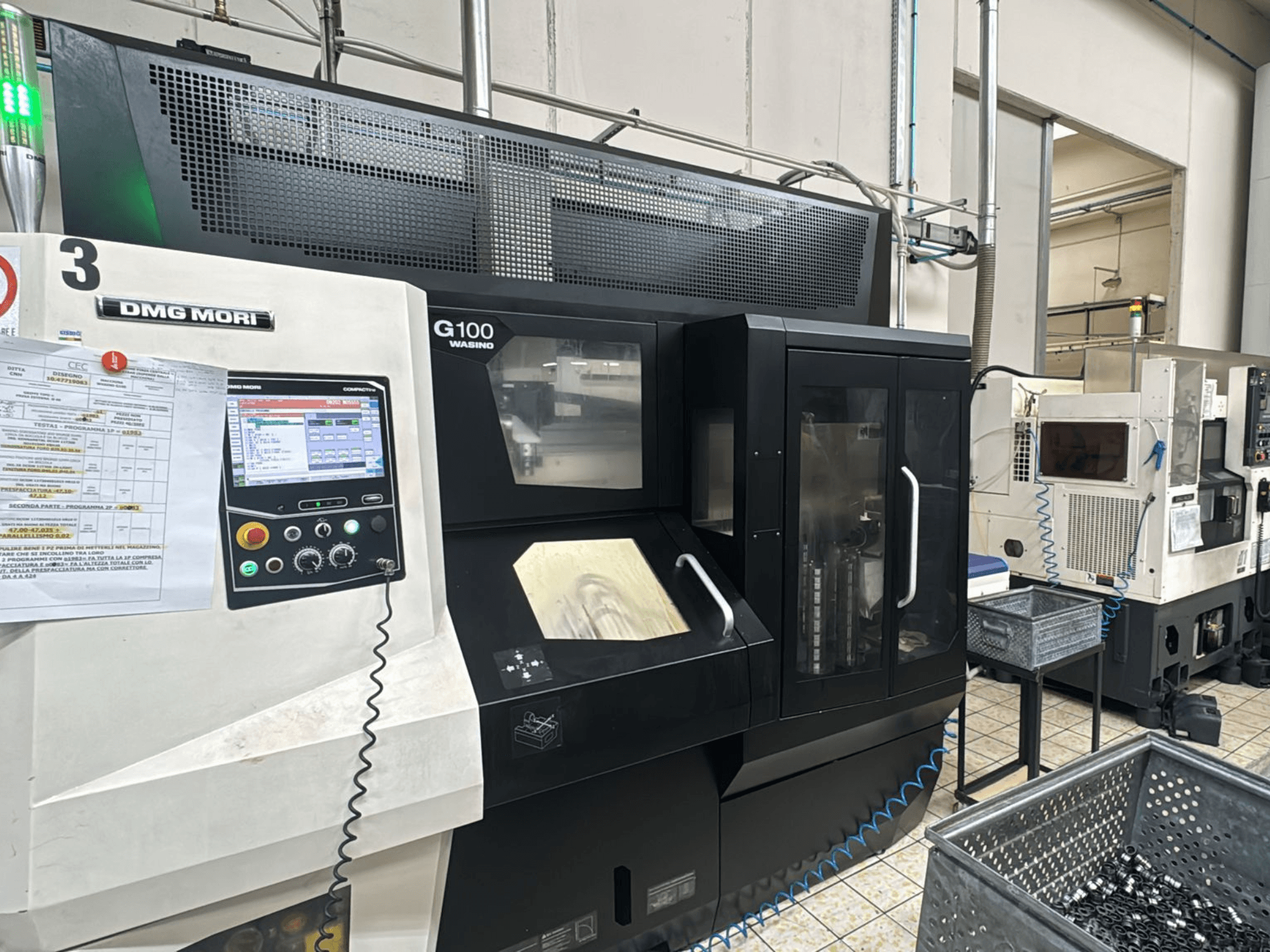 DMG MORI G100 staklės gamykloje, vaizdas iš priekio, su skaitmeniniu valdymo pultu ir šalia esančiomis įrankių talpyklomis.