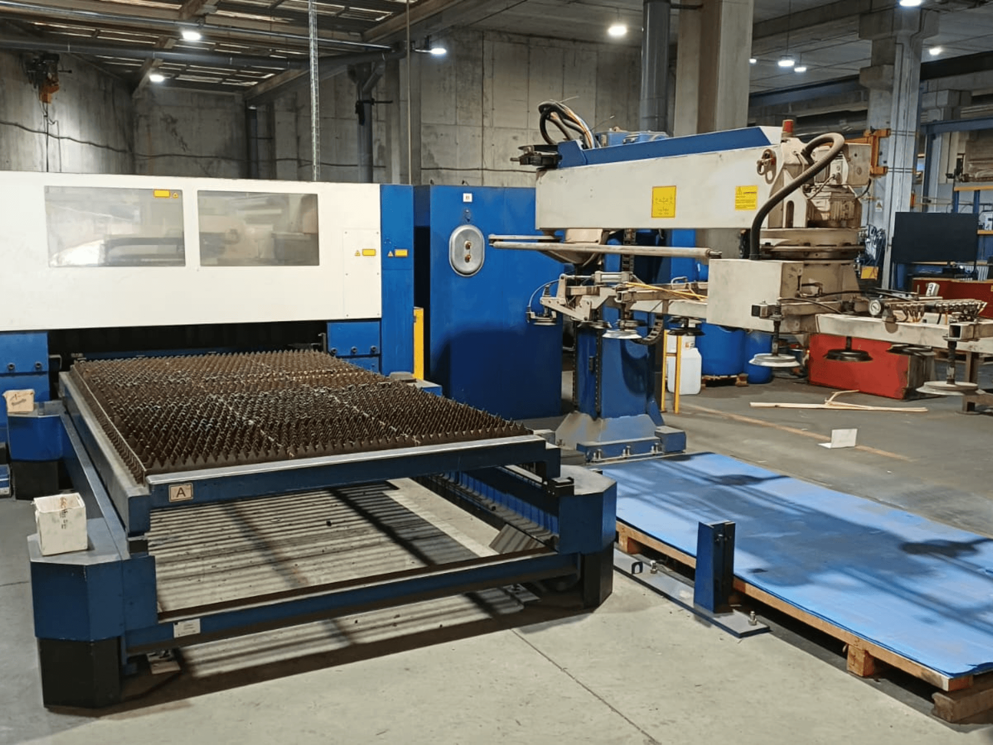 „TRUMPF TRUMATIC L 3030“ CO₂ lazerio pjovimo staklės, vaizdas iš šono, kuriame matomas pjovimo stalas su dygliuotu paviršiumi ir robotinė ranka.