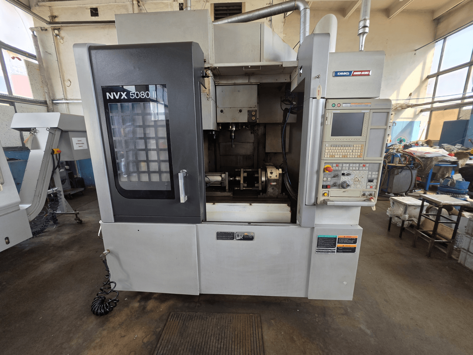 DMG Mori Seiki NVX 5080 II/40 vertikalaus apdirbimo centro vaizdas iš priekio su valdymo pultu ir permatomu langeliu, pro kurį matoma apdirbimo zona.