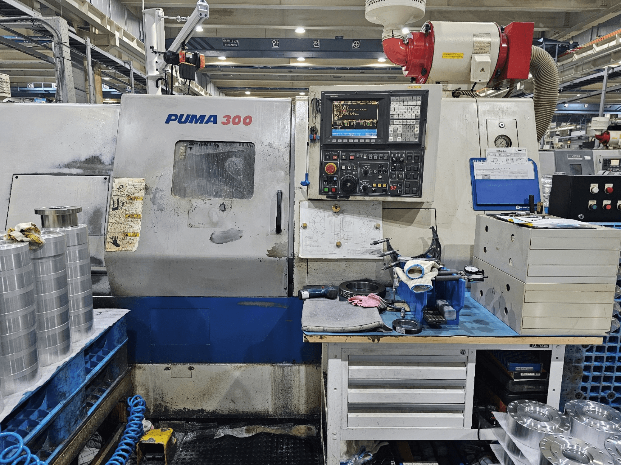PUMA 300 CNC staklės dirbtuvėse; vaizdas iš priekio, rodantis valdymo skydelį, įrankius ir metalines dalis ant darbo stalo.