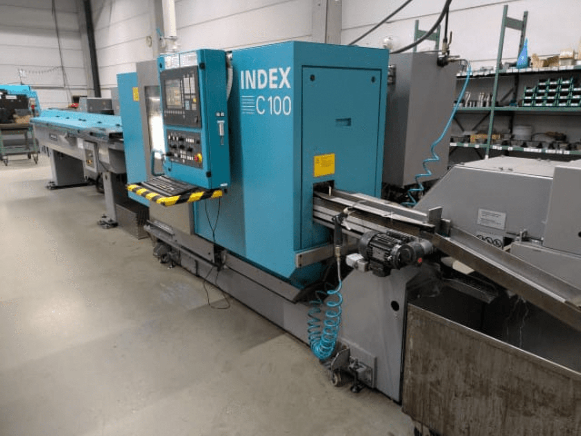 „Index C100“ horizontalios tekinimo staklės, vaizdas iš šono; su valdymo pultu, padavimo sistema ir metalo drožlių surinkimo dėže.