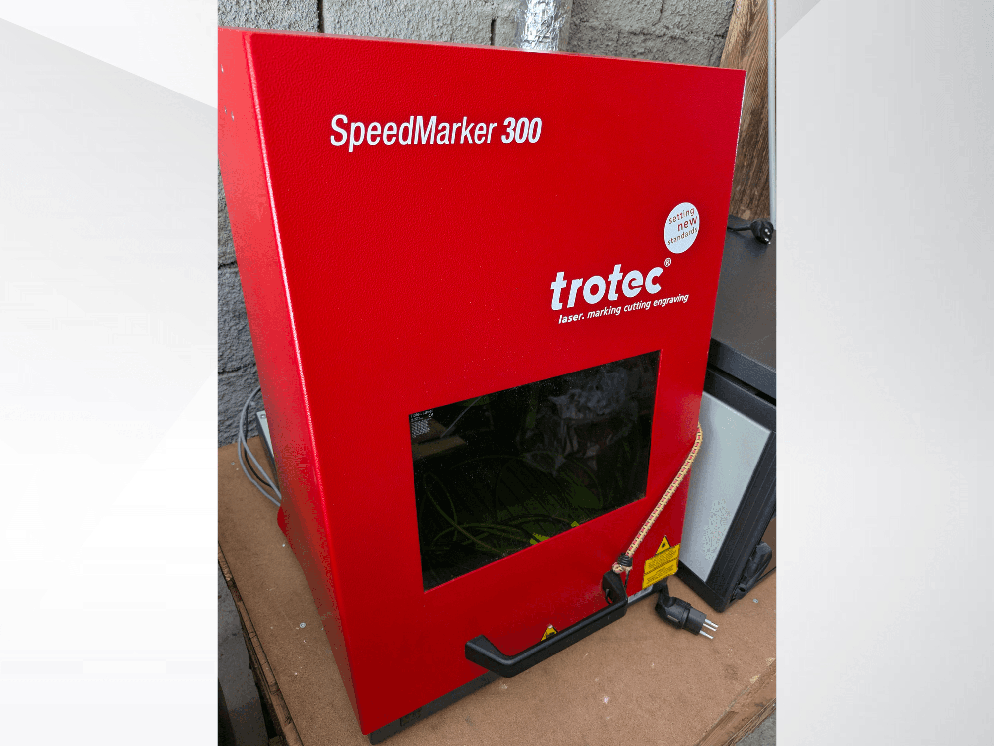 Raudonasis "Trotec SpeedMarker 300" lazerinis žymėjimo aparatas iš priekio, su dideliu stebėjimo langu ir maitinimo jungtimi.
