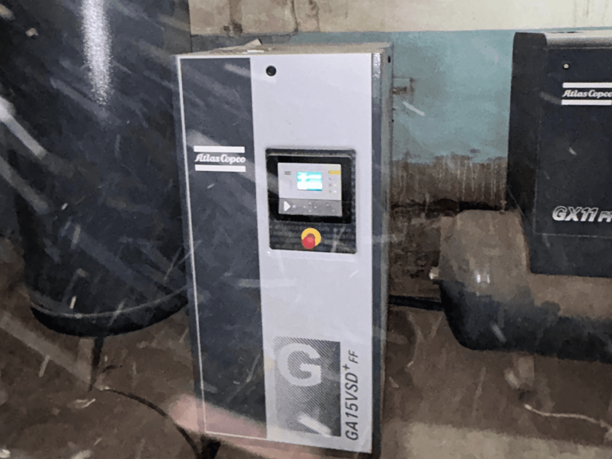 „Atlas Copco“ GA15VSD+FF oro kompresorius, vaizdas iš priekio; matomas valdymo skydelis su ekranu ir raudonu avarinio sustabdymo mygtuku.
