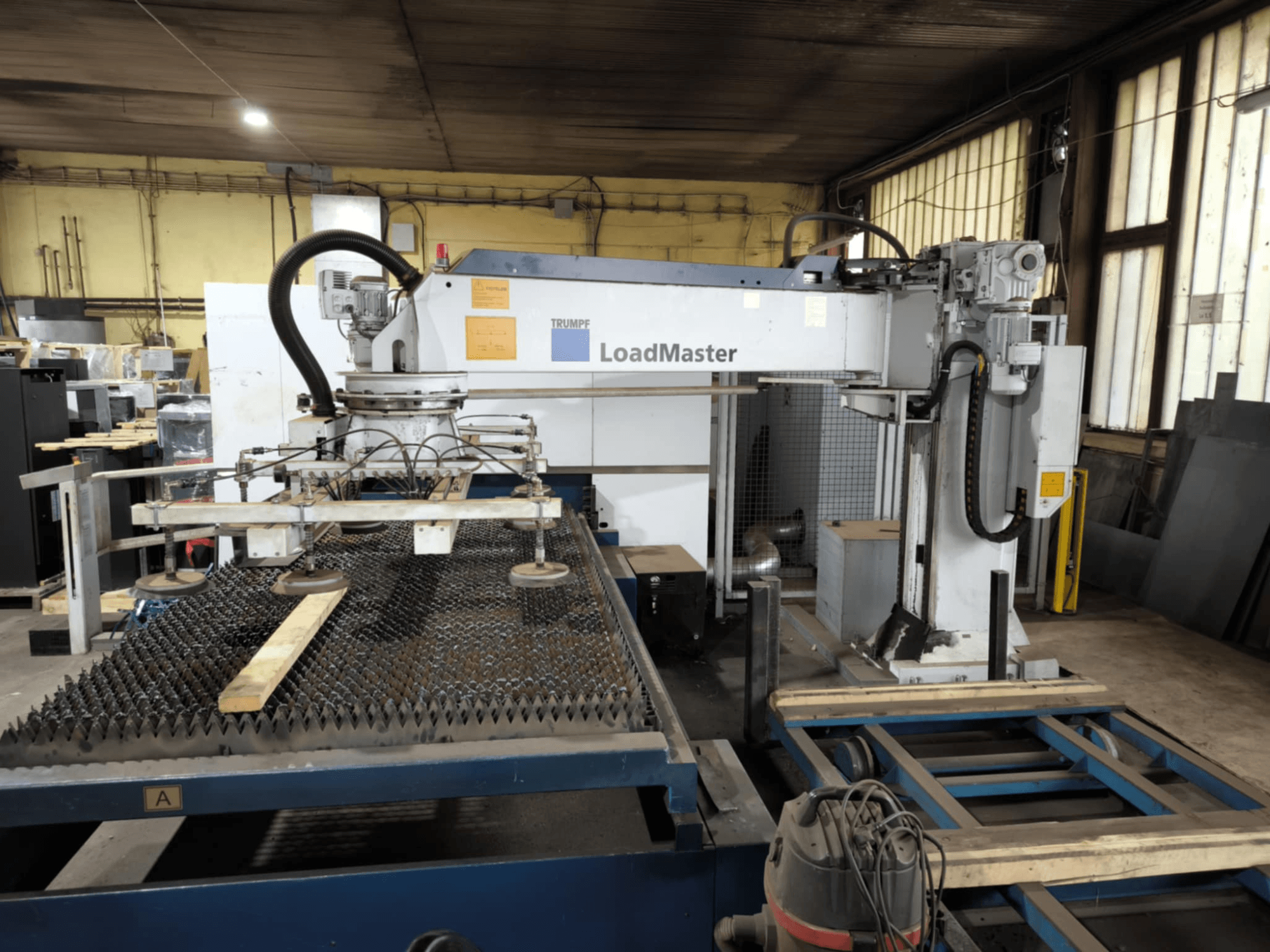 TRUMPF TRULASER 3030 FIBER pjaustymo staklės, vaizdas iš priekio, su "LoadMaster" sistema ir ant grotelėmis padengto darbo stalo.