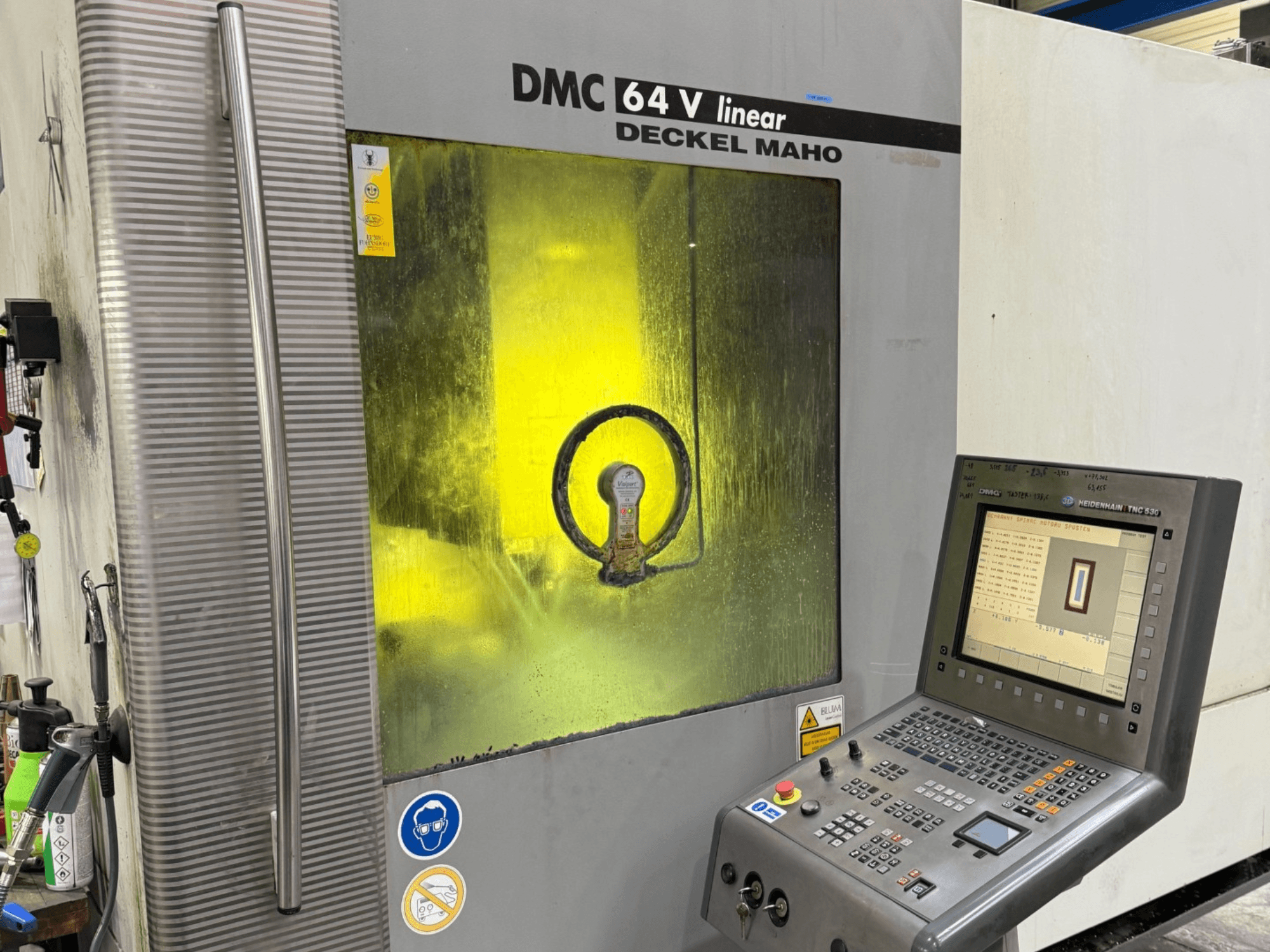 Deckel Maho DMC 64 V linear, mašinos vaizdas iš priekio su valdymo skydeliu ir geltonai tonuotu langeliu, pro kurį matomi viduje esantys mechanizmai.