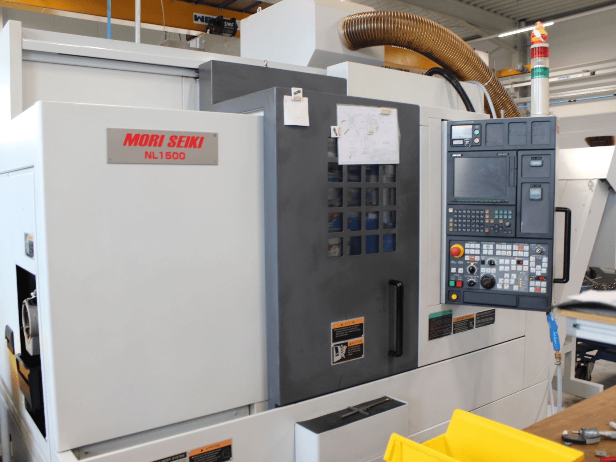MORI SEIKI NL 1500 SY tekinimo-frezavimo staklių šoninis vaizdas, kuriame matomas valdymo pultas, darbo saugos etiketės ir aušinimo sistema.