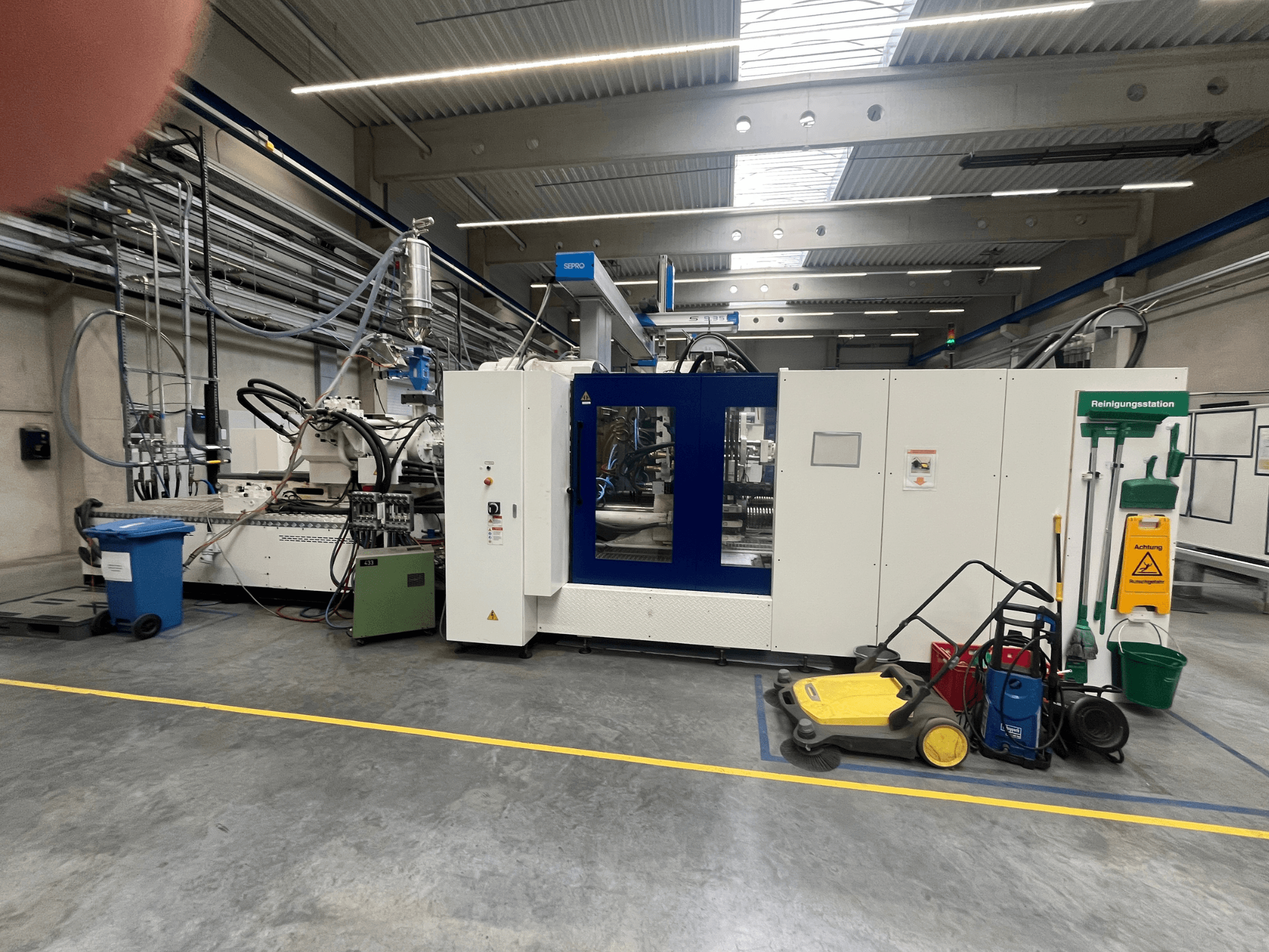 Pramoninė mašina gamykloje, vaizdas iš priekio, šalia mėlynos ir baltos plokštės, žarnos ir valymo įranga.
