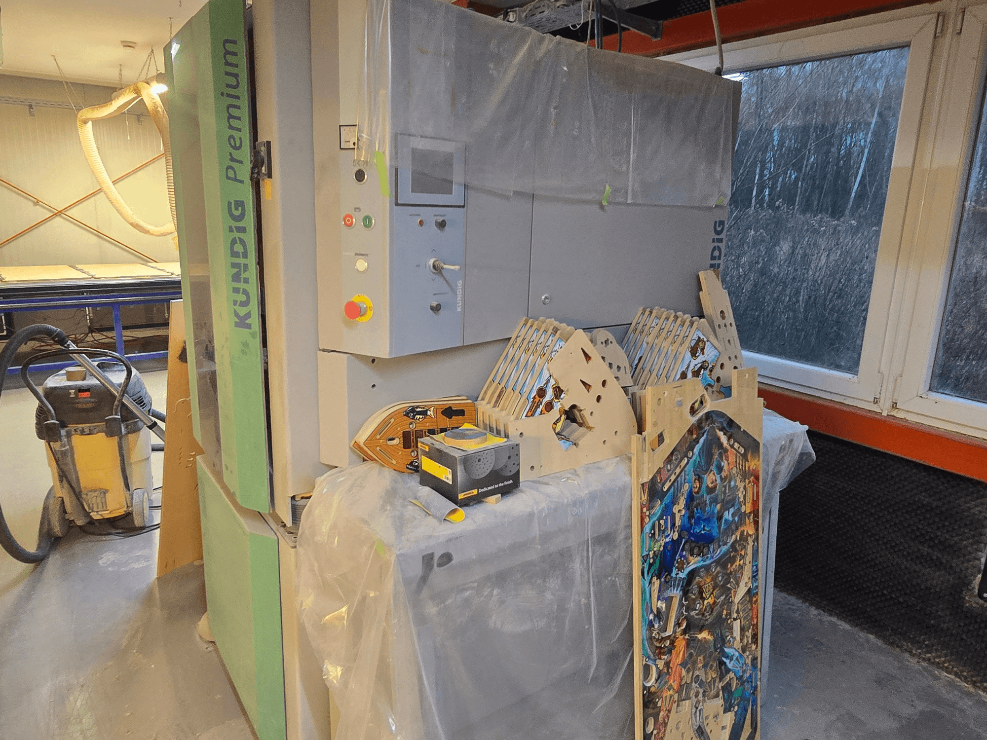 Juostinis šlifuoklis "Kundig Premium-1 1100-C" dirbtuvėse, matomas valdymo skydelis ir medienos dalys, šoninis vaizdas, fone - dulkių siurblys.