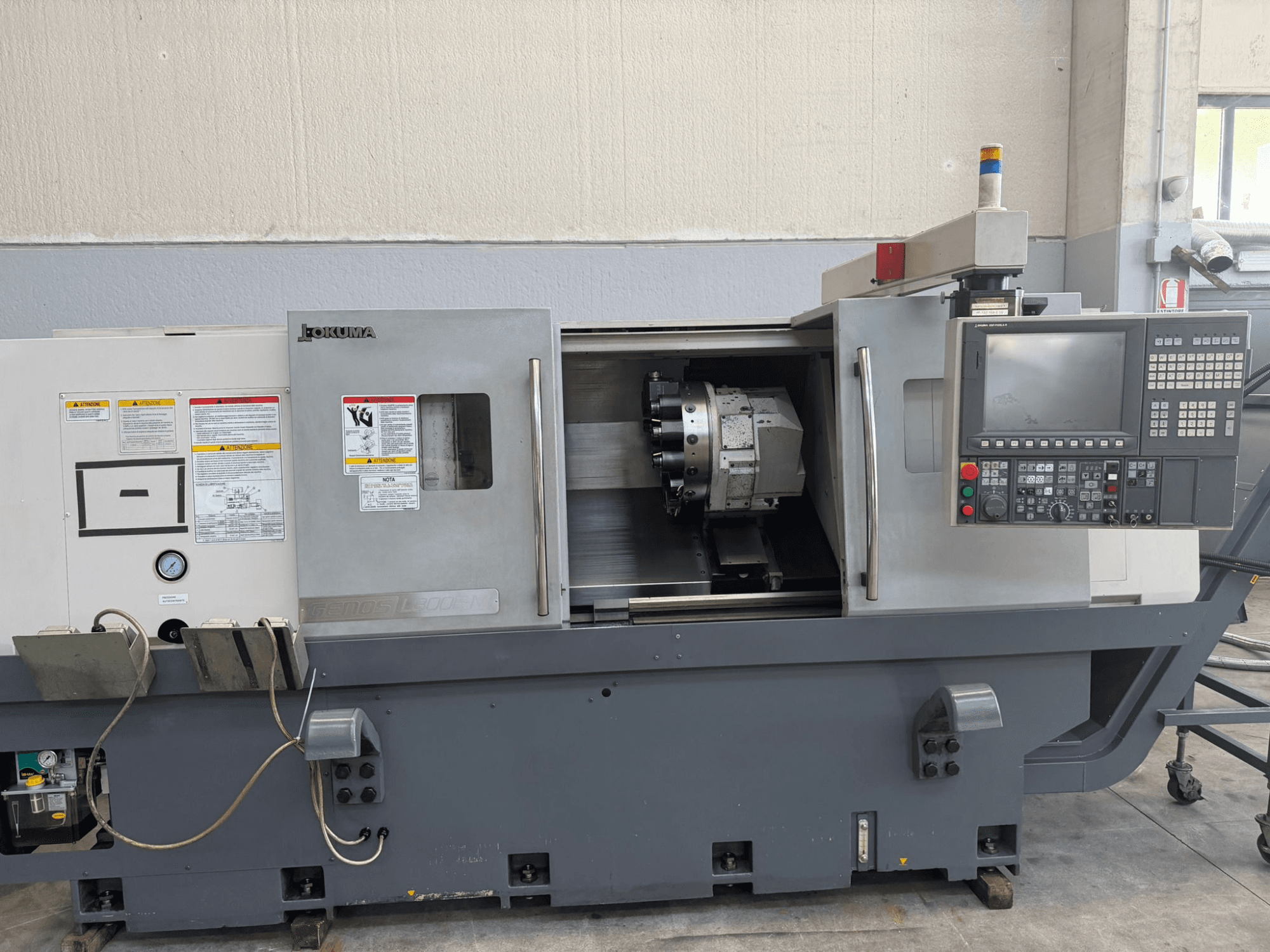 "Okuma" CNC tekinimo staklės, priekinis vaizdas, kuriame matomas valdymo skydelis, pjovimo įrankio laikiklis ir saugos įspėjimai šone.