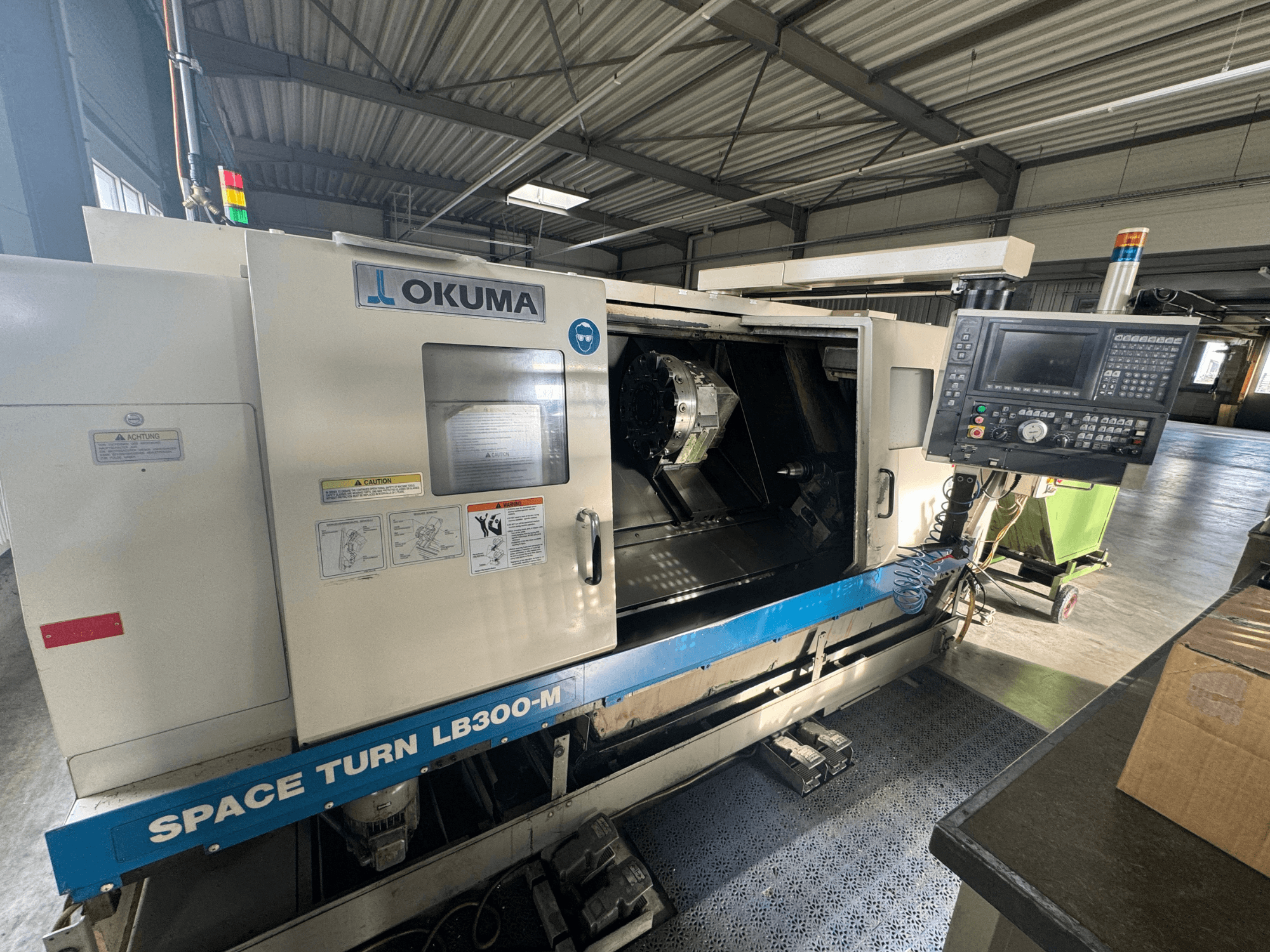 Okuma Space Turn LB300-M CNC staklės iš priekio, su valdymo skydeliu ir atviromis durelėmis, atveriančiomis vidinę apdirbimo zoną.