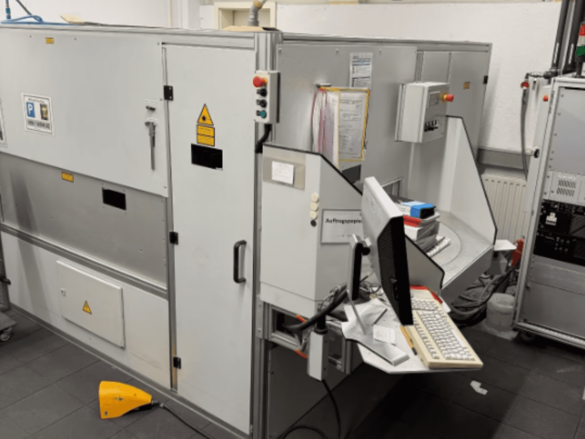 Pramoninė mašina su kompiuterio terminalu, vaizdas iš šono; yra valdymo mygtukai ir įspėjamieji saugos ženklai.