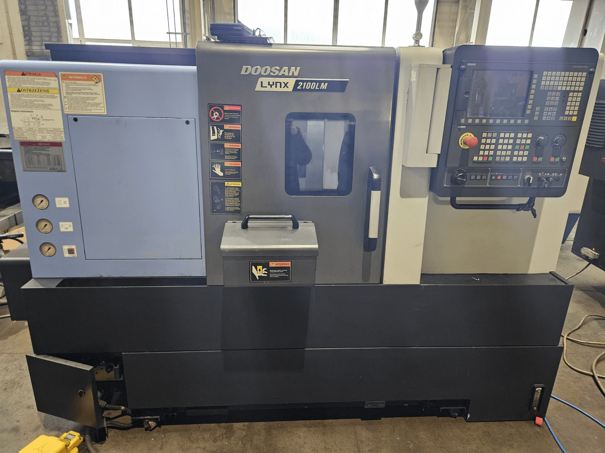 CNC staklės "Doosan Lynx 2100LM", vaizdas iš priekio, matomas valdymo skydelis, oro slėgio matuokliai ir saugos etiketės.