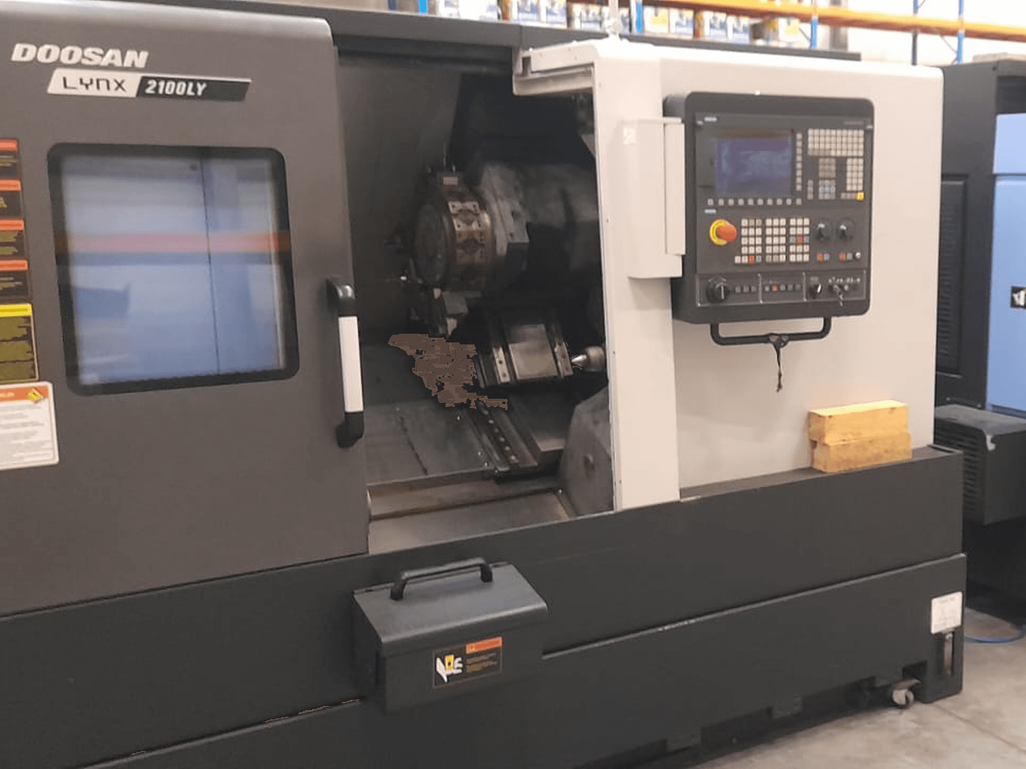 CNC tekinimo staklės "Doosan Lynx 2100LY", priekinis vaizdas, kuriame matyti valdymo skydelis, įrankių laikiklis ir vidinė darbo vieta; matomos techninės etiketės.