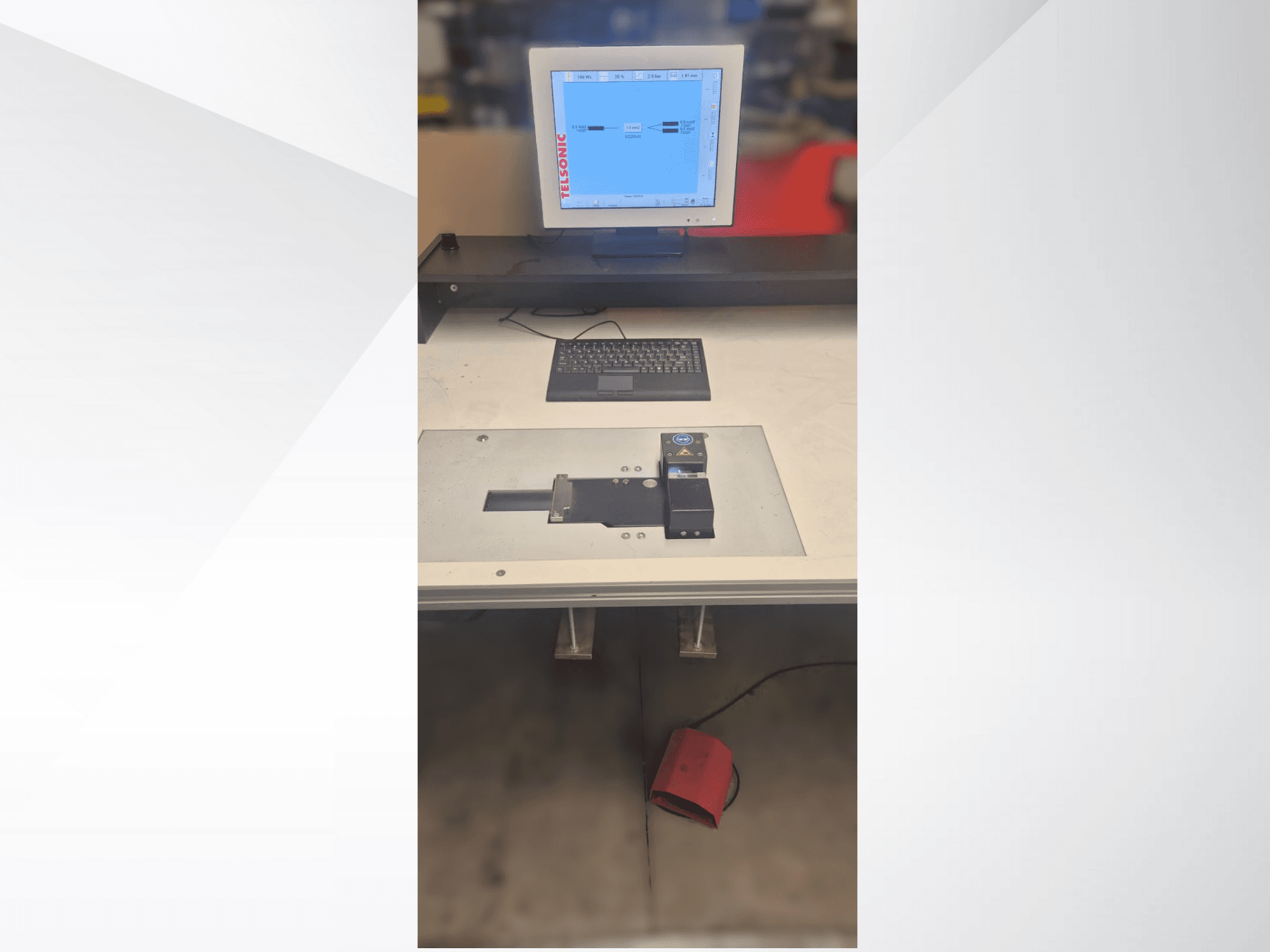 Iš viršaus į apačią nuleistas testavimo stoties vaizdas su kompiuterio monitoriumi, klaviatūra ir valdymo pultu, kuriame yra mygtukas ir žalia lemputė.