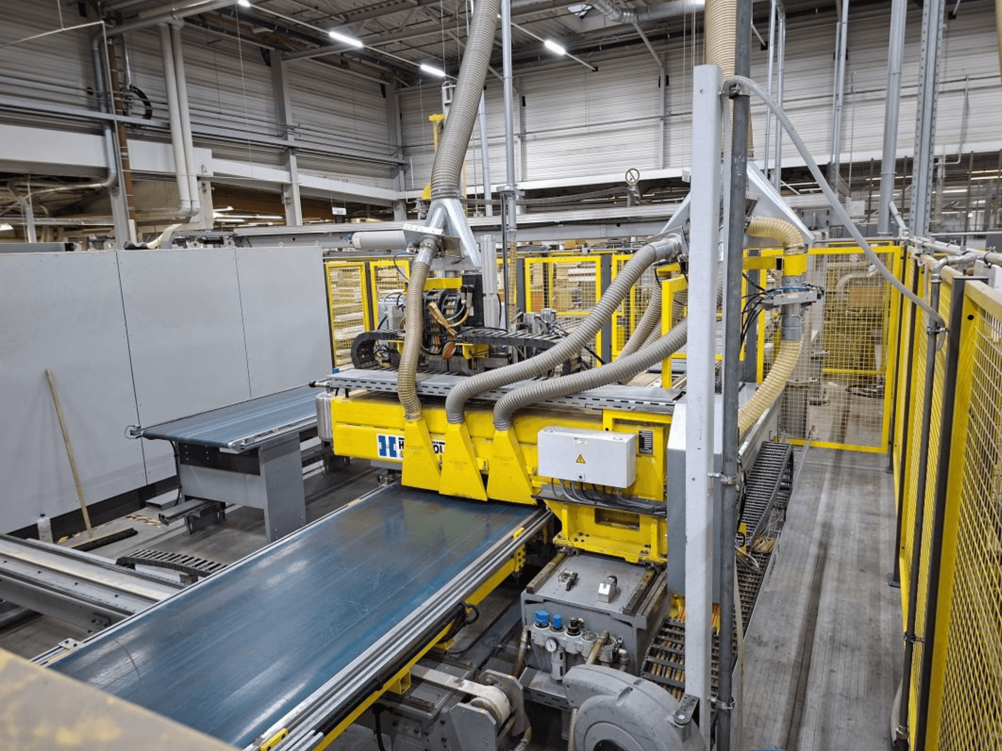 Pramoninė konvejerių sistema iš šono, kurioje matomos geltonos spalvos mašinos, valdymo pultas ir gamybai skirtos žarnos.