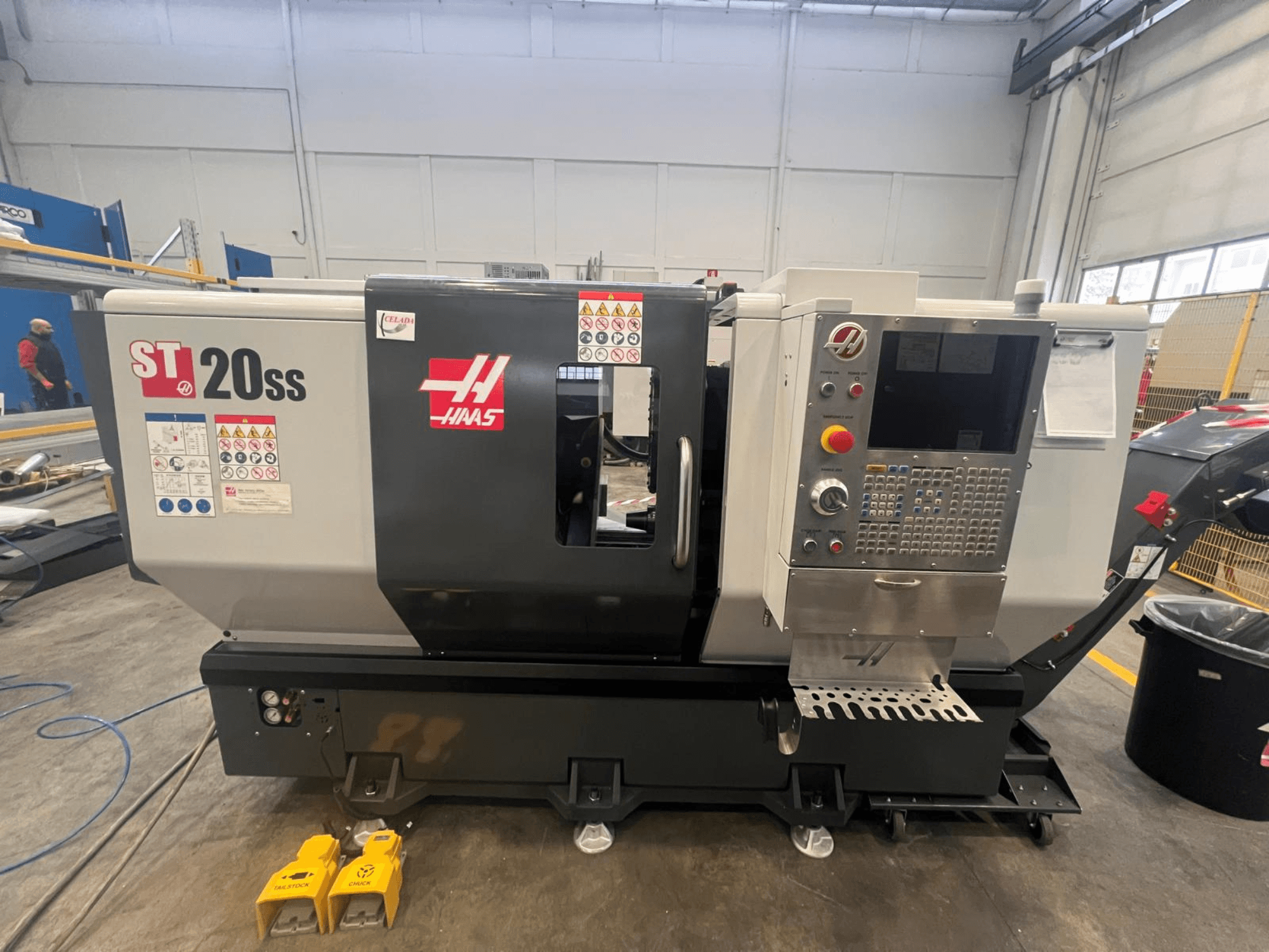 Haas ST 20SS CNC tekinimo staklės iš šono, su valdymo skydeliu, saugos etiketėmis ir ant grindų esančiais kojiniais pedalais.