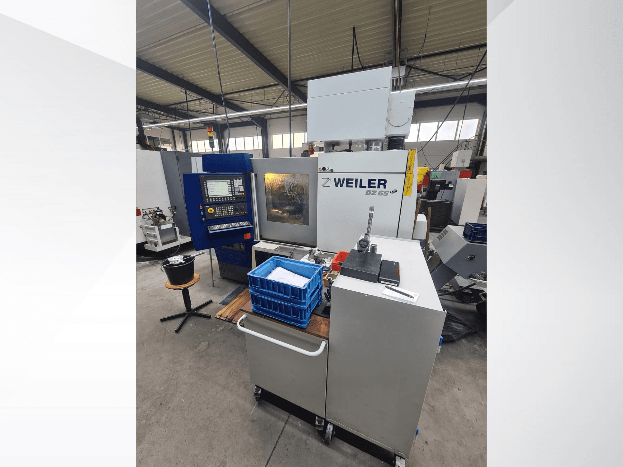 Weiler DZ 65 CNC staklės dirbtuvėse, vaizdas iš priekio, matomas valdymo skydelis, įrankiai ir mėlynos sandėliavimo dėžės.