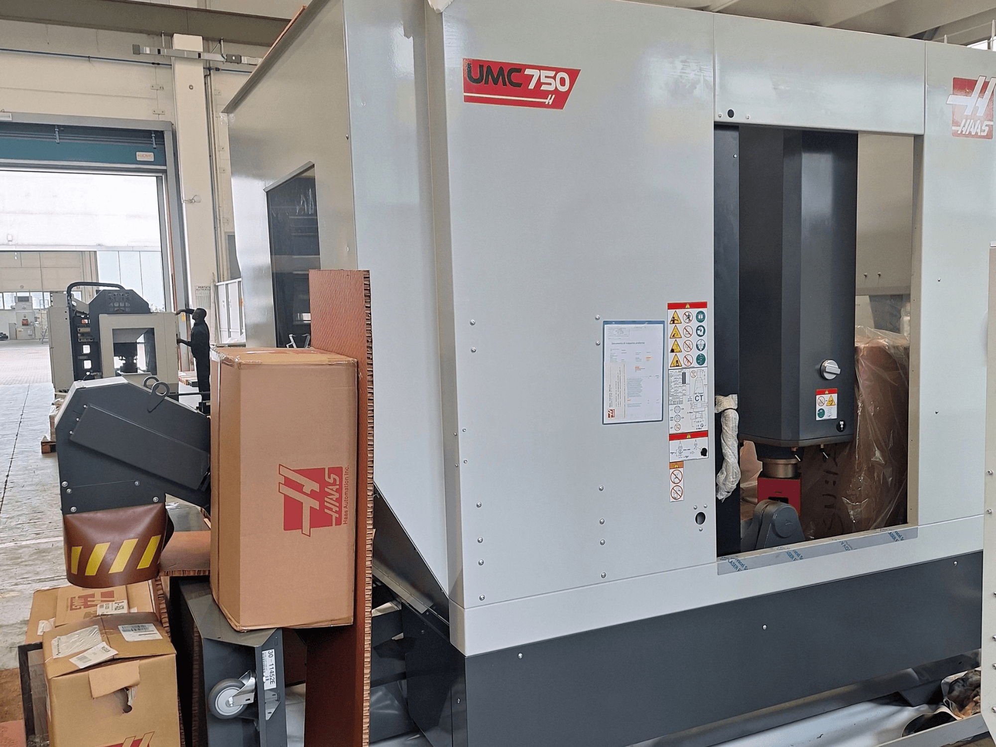 Universalus apdirbimo centras „HAAS UMC-750“, vaizdas iš šono, kuriame matomas valdymo skydas ir įrankių laikymo vieta su įspėjamosiomis etiketėmis bei pakuotėmis.