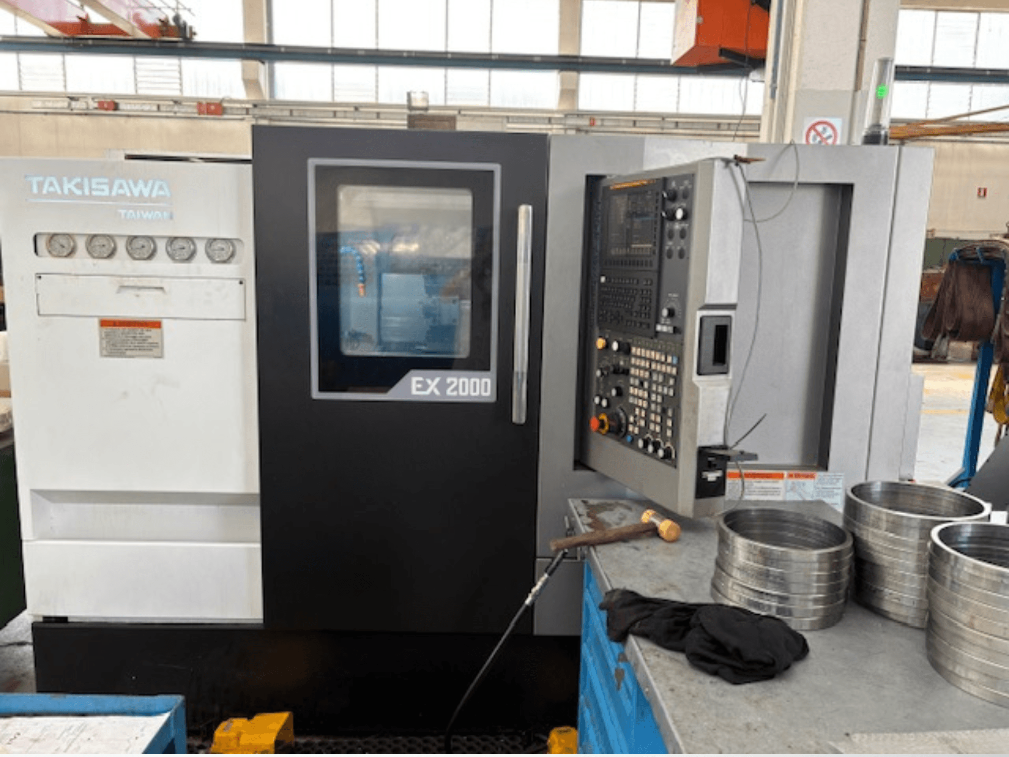 CNC staklės "Takisawa EX 2000", vaizdas iš priekio, valdymo skydelis, matuokliai ir metalinės detalės ant dirbtuvių stalo.