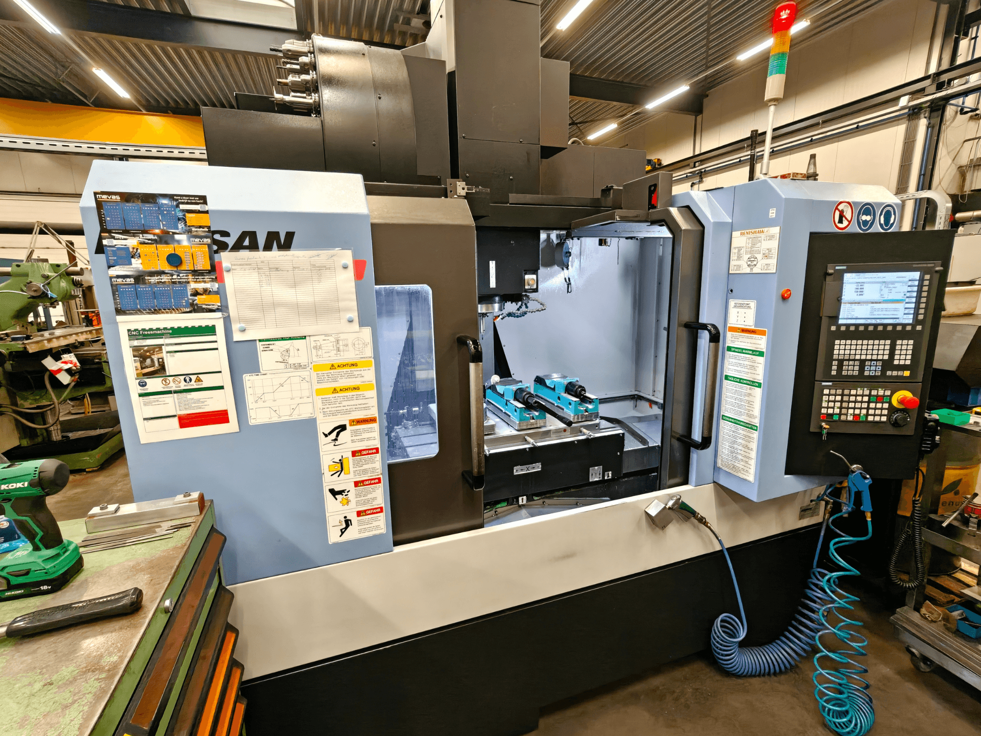 DOOSAN DNM 400 II vertikalusis apdirbimo centras dirbtuvėse, su valdikliais, įrankių laikikliais ir saugos ženklais, vaizdas iš priekio.