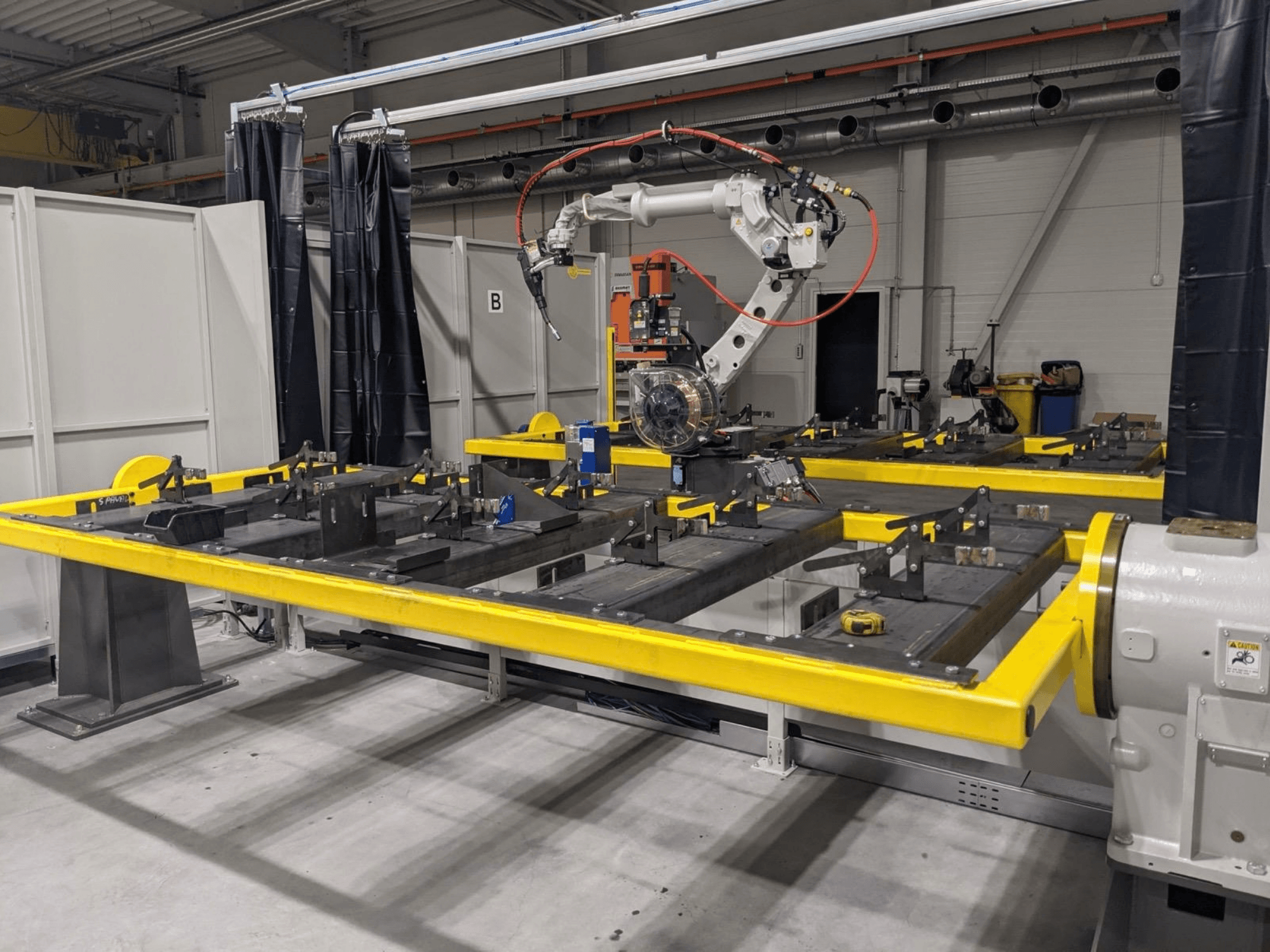 „PANASONIC TAWERS TM-2000WG3“ robotinės rankos vaizdas iš priekio; ji įtaisyta virš geltono darbo stalo, ant kurio guli surinkimui skirtos metalinės detalės.