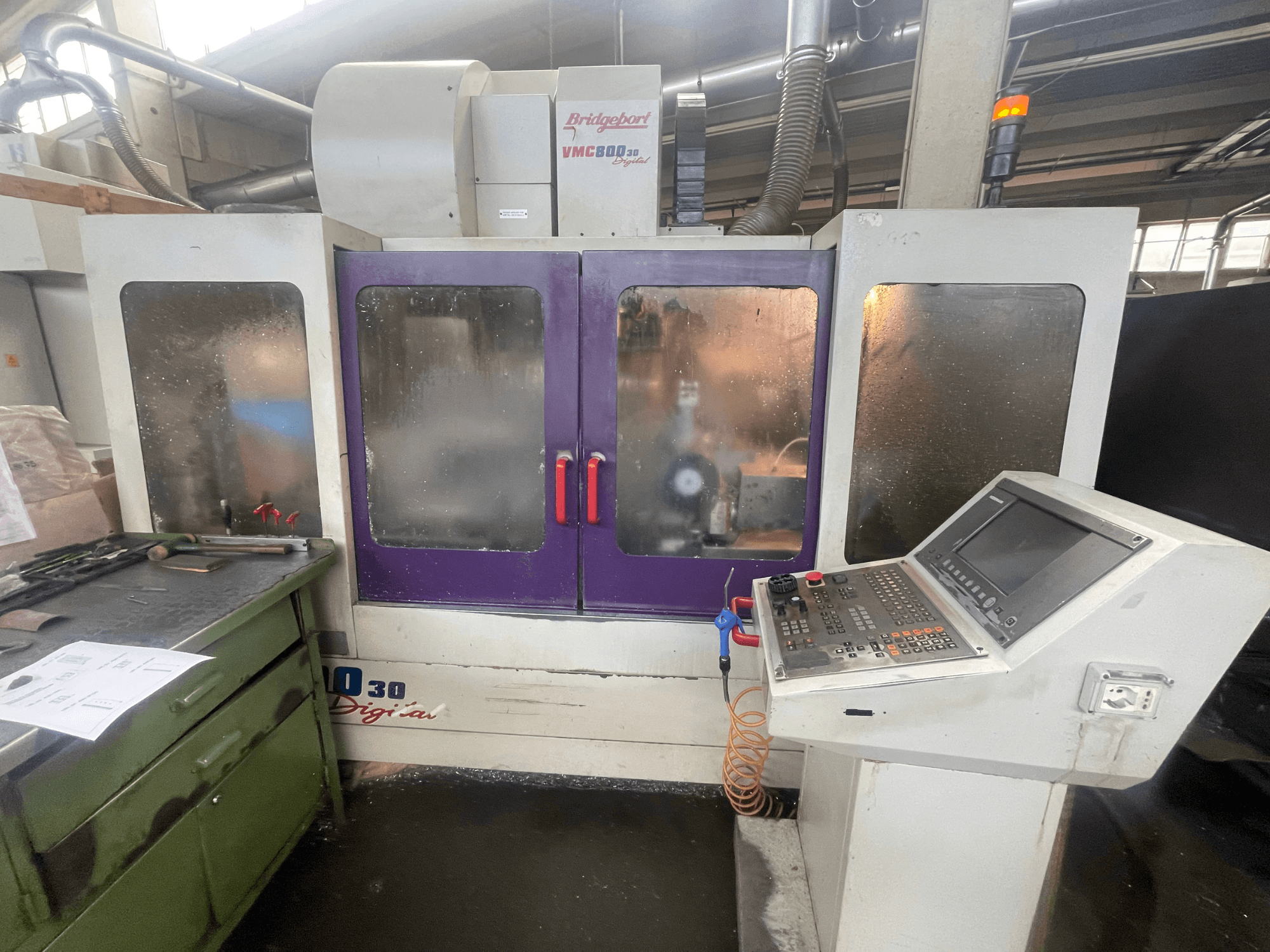 Vertikalusis apdirbimo centras „Bridgeport VMC 800/30“, vaizdas iš priekio, kuriame matomas valdymo pultas ir violetinės spalvos durelės, o ant greta esančio paviršiaus – įrankiai.
