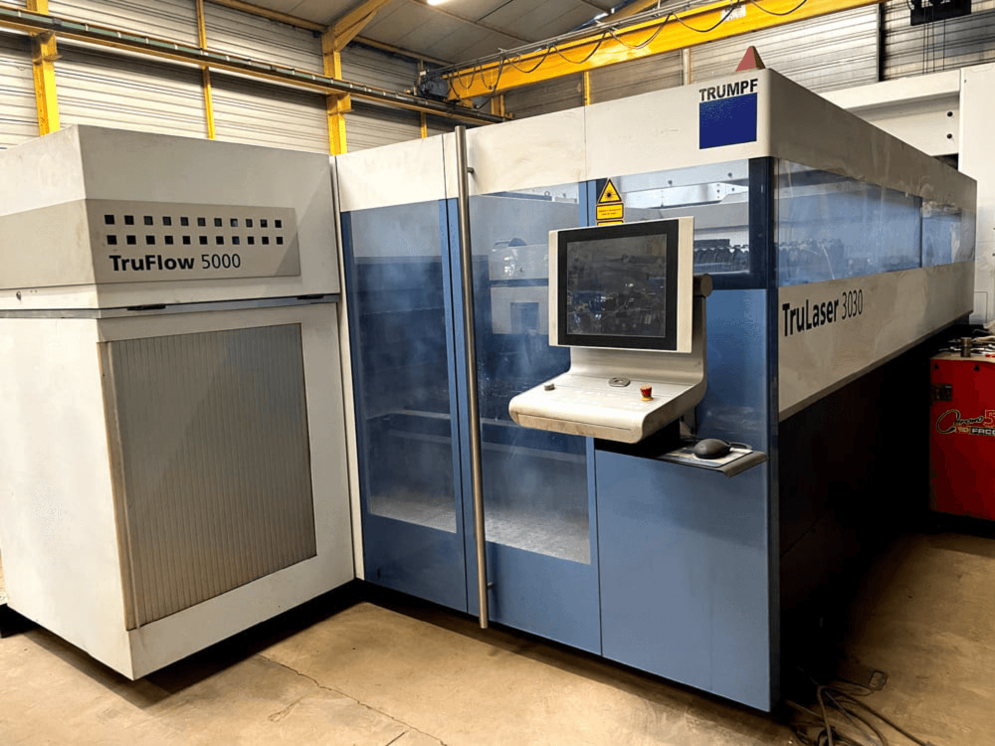 „TRUMPF TruLaser 3030“ CO₂ lazerinio pjovimo staklės, vaizdas iš priekio, su valdymo pultu ir šalia esančiu „TruFlow 5000“ įrenginiu.