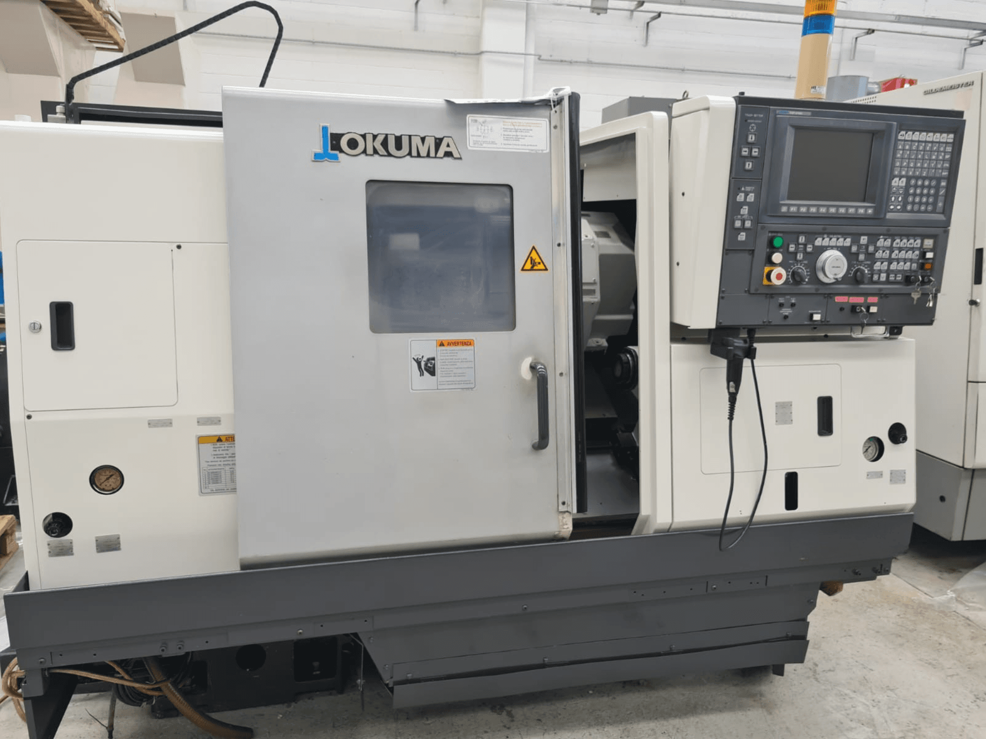 Okuma LB 300M X 500 horizontaliosios tekinimo staklės iš šono, matyti valdymo skydelis, prieiga prie suklio ir saugos įspėjimai.